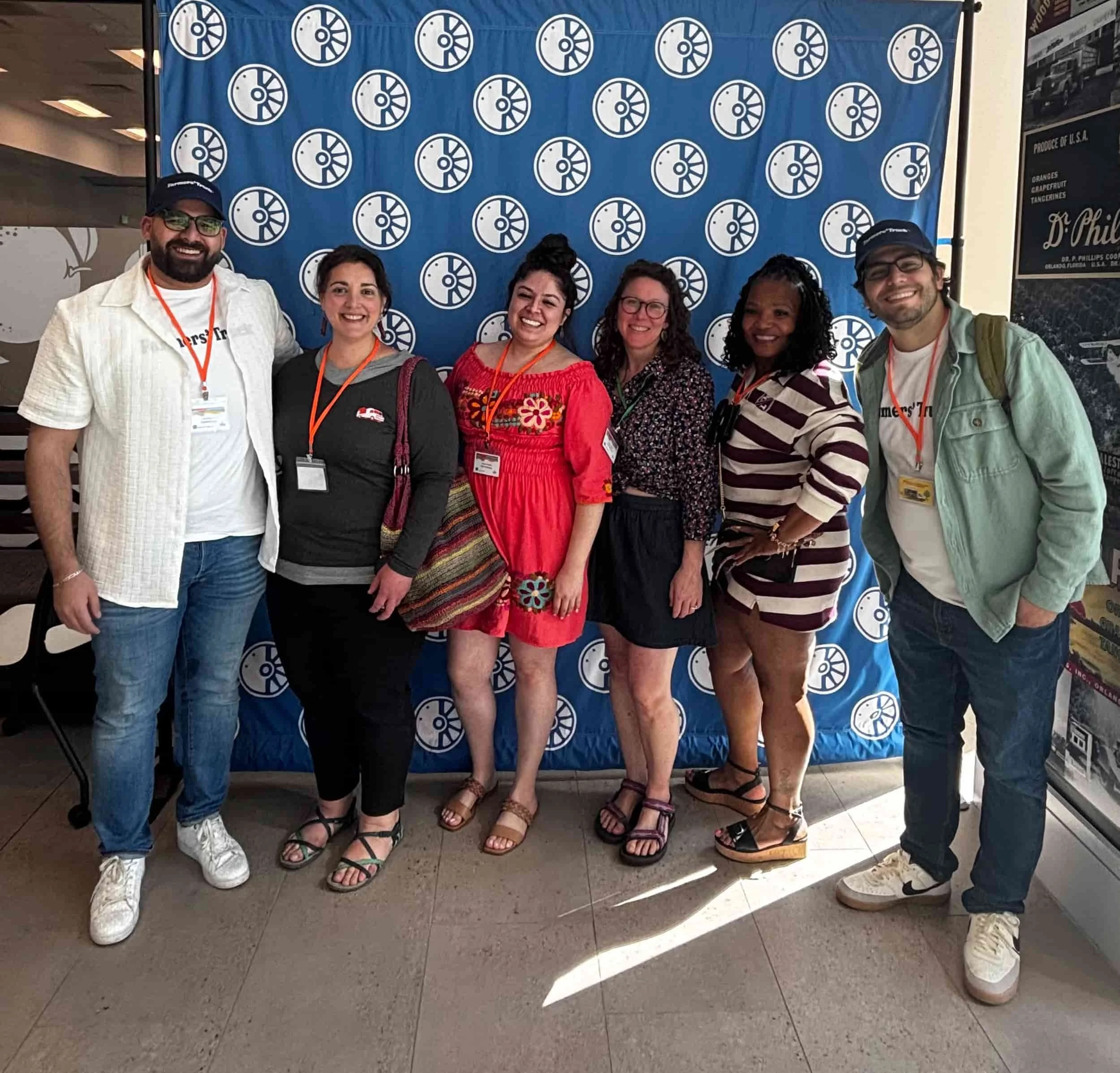 Group of six smiling people standing in front of a blue backdrop with white circular logos, inside a building. They are wearing casual and semi-casual outfits with event badges around their necks.