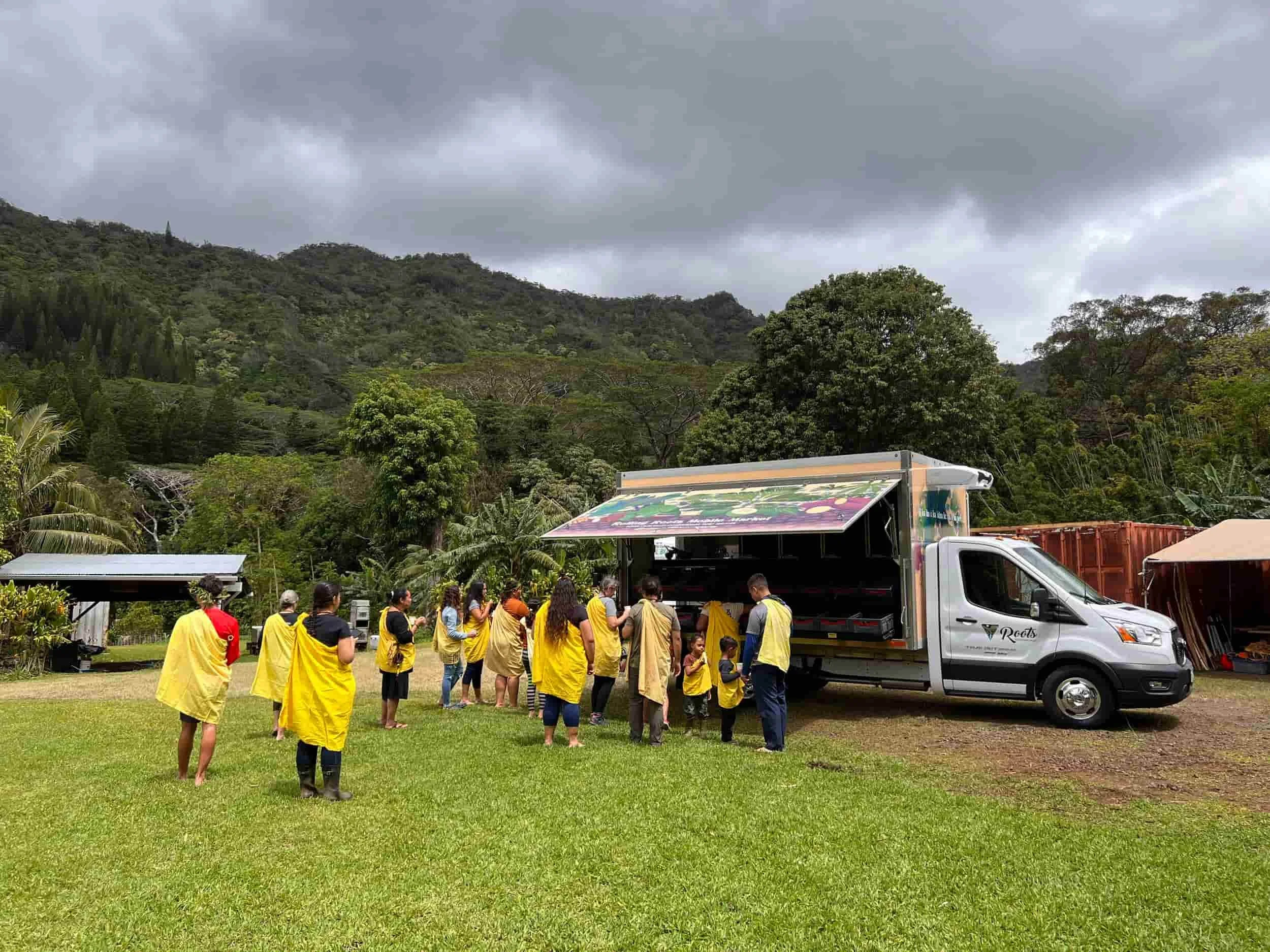 People in yellow cloaks standing in line outside a mobile food truck in a green, hilly landscape with trees and dark cloudy sky.