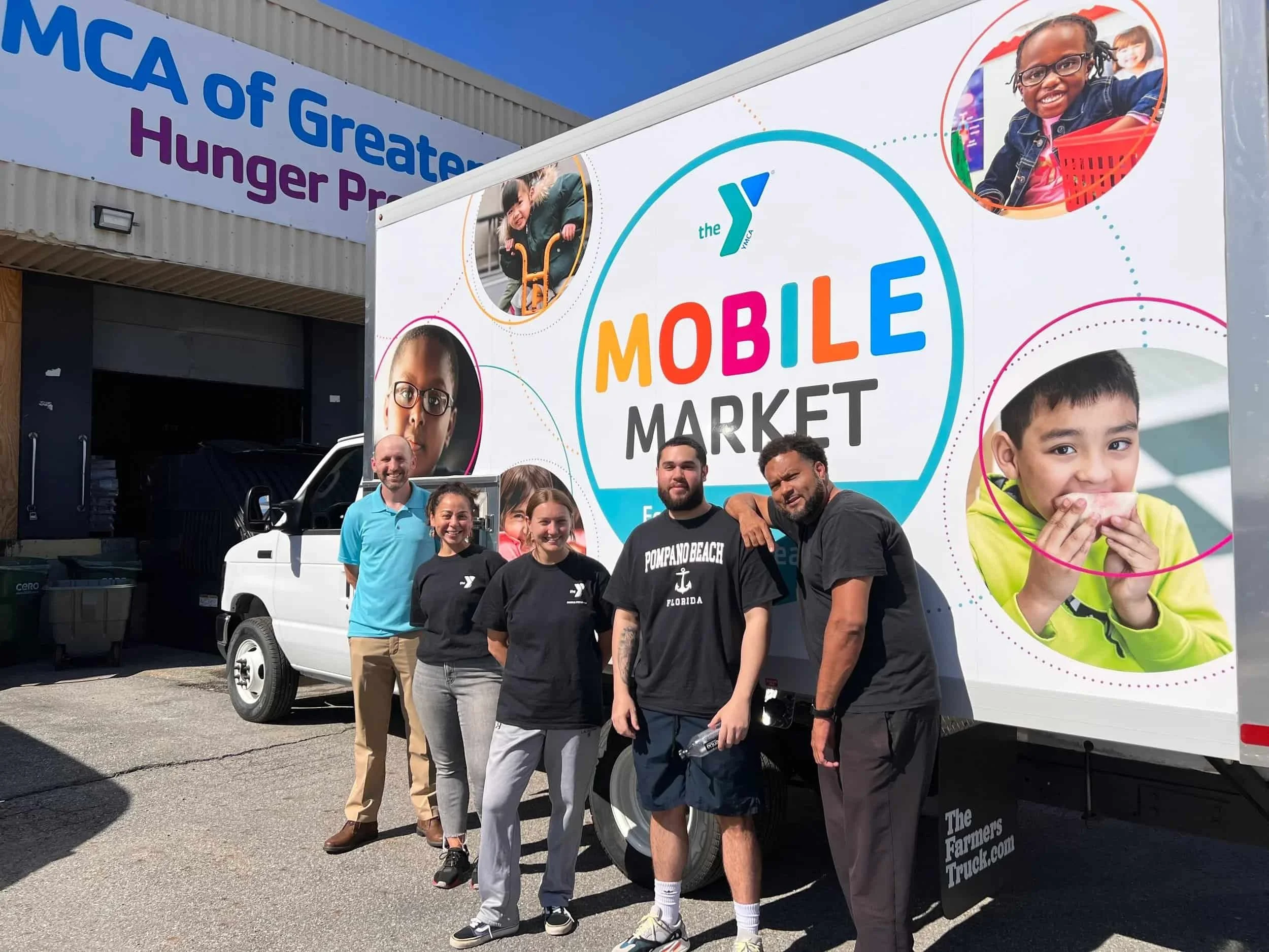 Group of five people standing in front of a large Y-shaped printed truck with a sign that reads 'Mobile Market' and features photos of children and families; background shows a building with a sign for 'MCA of Greater Hunger P'. it is sunny and clear