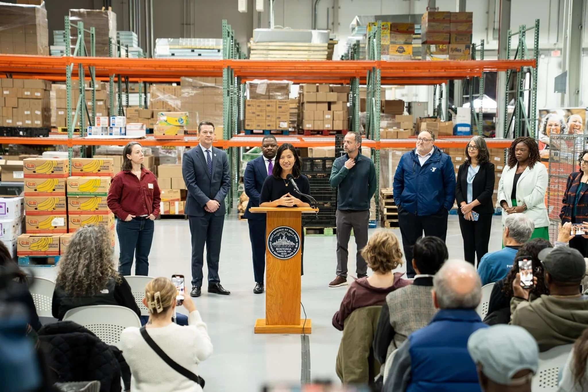 A woman speaking at a podium during a press conference in a warehouse, with several individuals standing behind her and people sitting in chairs listening, some taking photos.