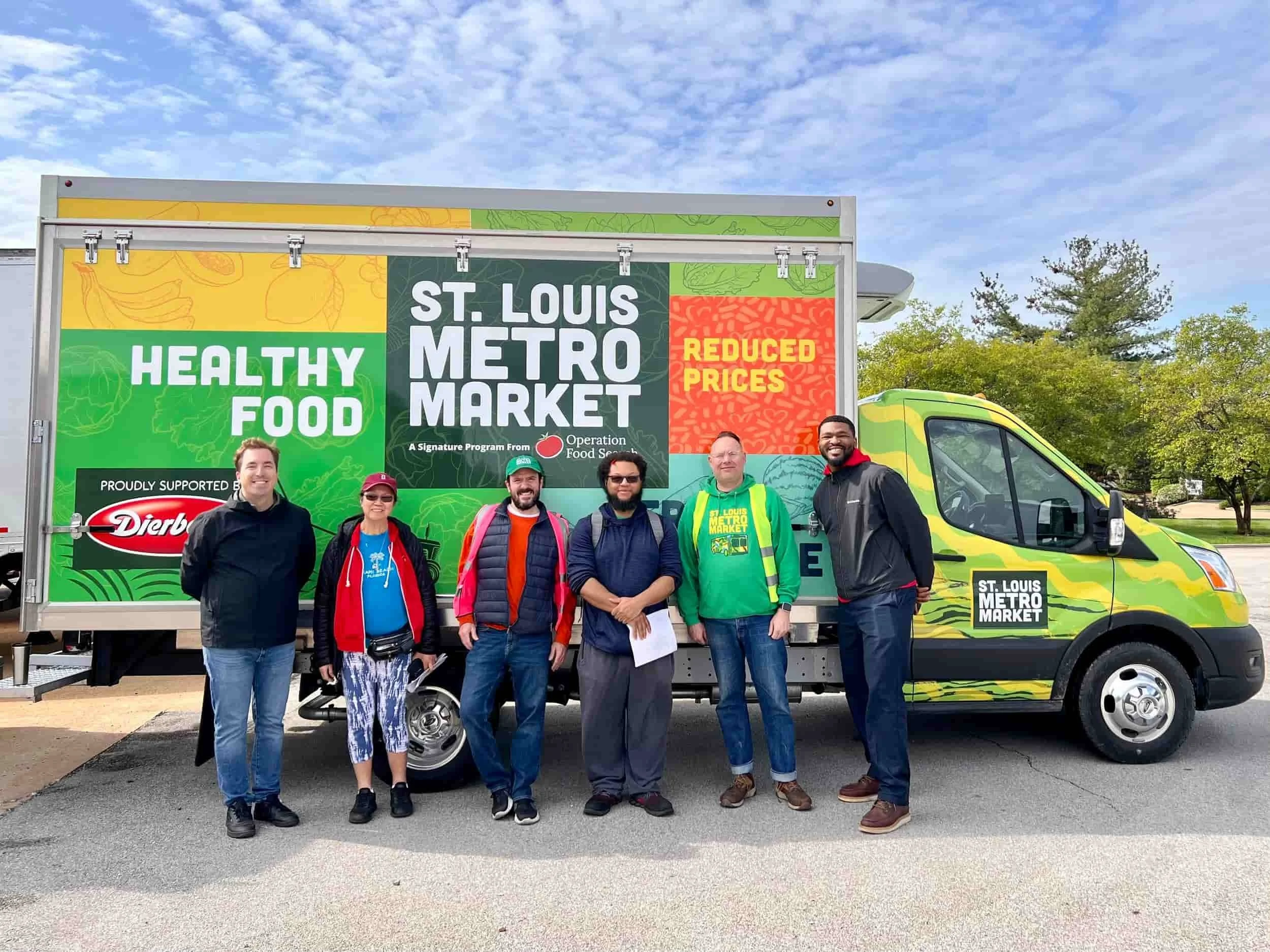 Group of six people standing in front of a mobile health food market truck with a colorful sign that reads 'Healthy Food', 'St. Louis Metro Market', and 'Reduced Prices'. The truck has a green and yellow camouflage pattern, and the people are smiling