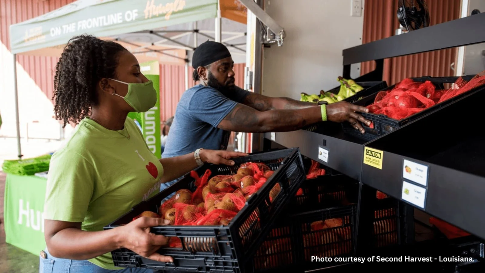 A woman and a man are selecting apples from a produce shelf at a farmers market. The woman is wearing a green face mask and a lime green shirt, while the man is wearing a black head covering and a blue shirt. They are placing apples into black basket