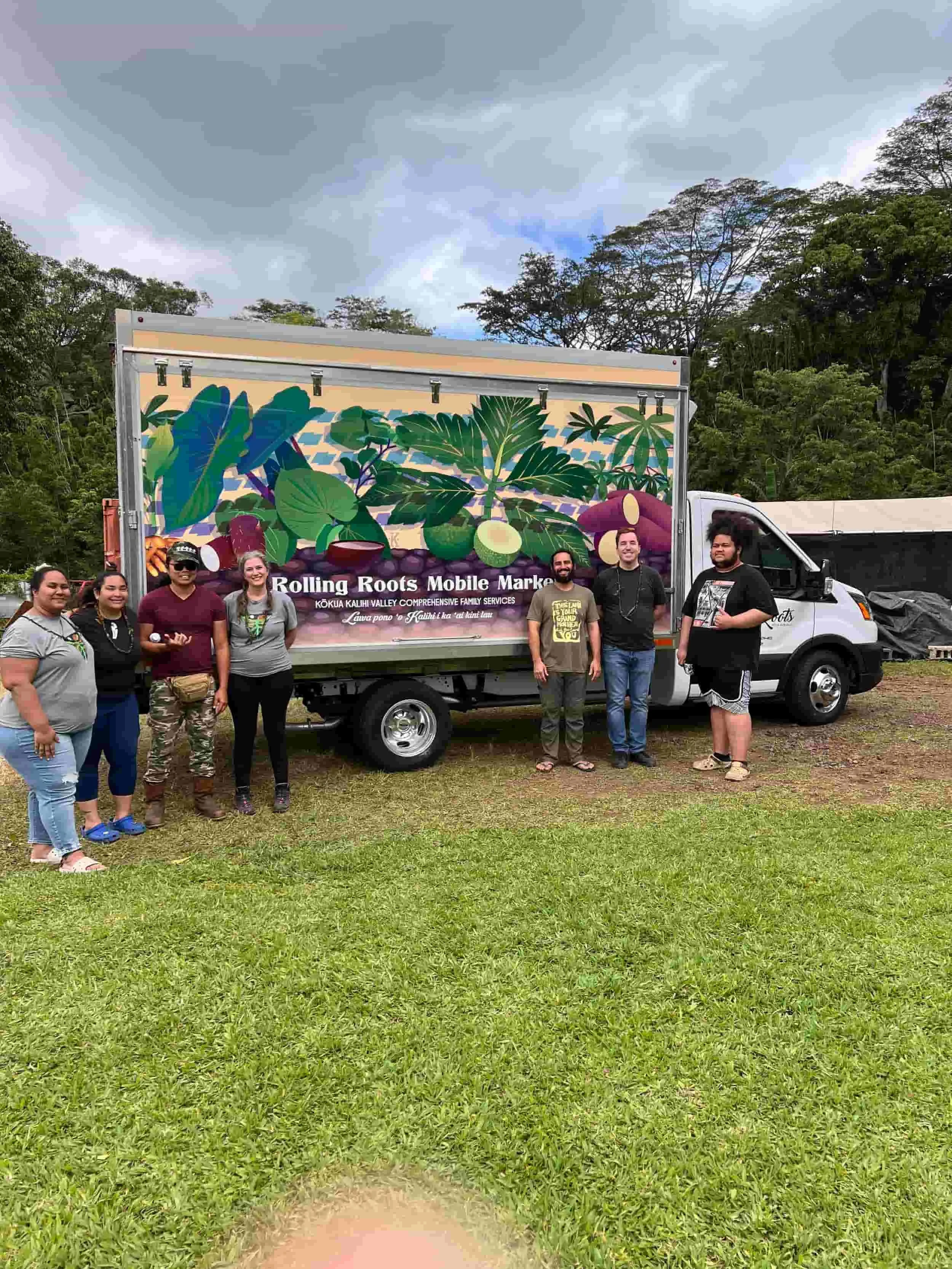 A group of seven people standing in front of a truck with a colorful graphic and the text 'Rolling Roots Mobile Market' and 'Kōkua Kālhi Valley Comprehensive Family Services' on it, outdoors on grassy terrain with trees and cloudy sky in the backgrou