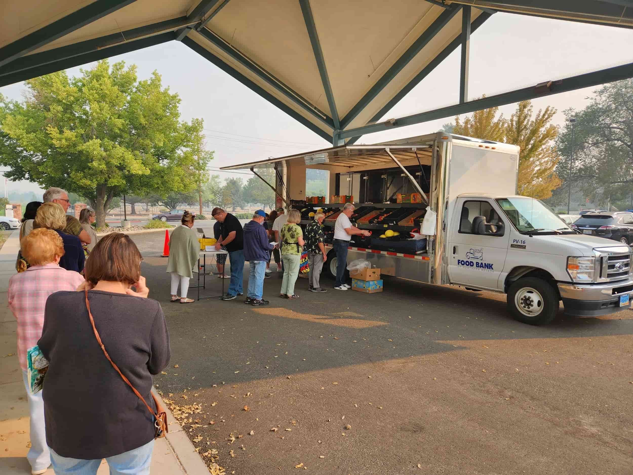 People lining up outside a food bank truck receiving groceries in an outdoor parking lot with trees and a building in the background.