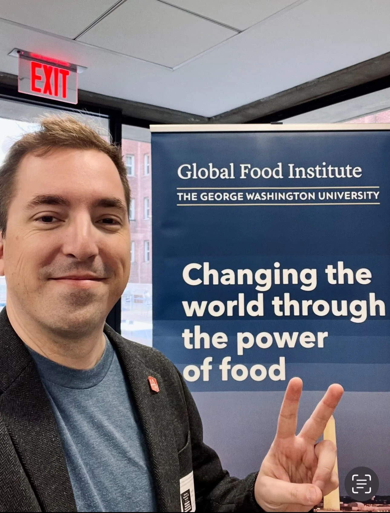A man smiling and showing a peace sign in front of a blue banner for the Global Food Institute at George Washington University, with an EXIT sign above.