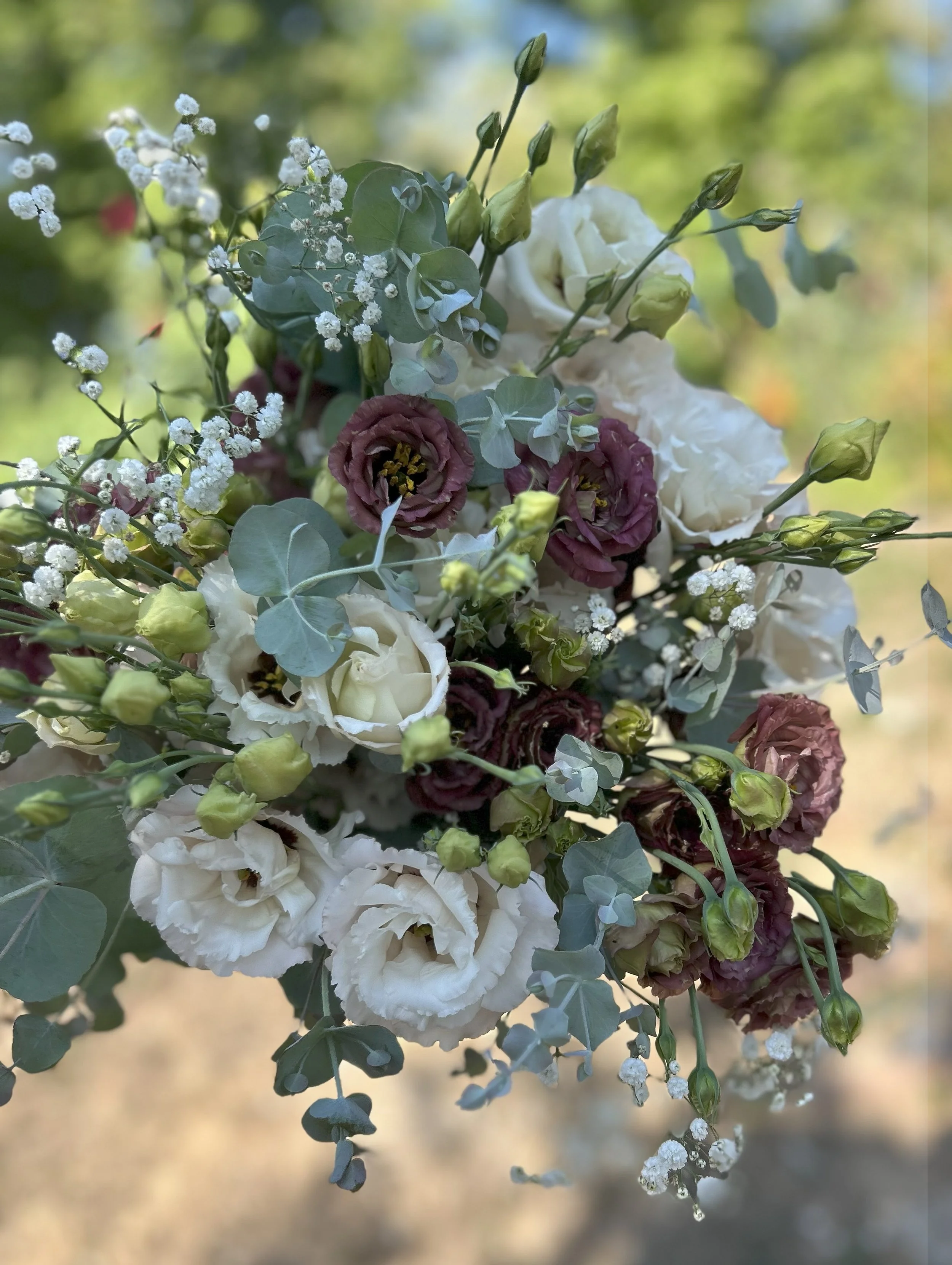 A close-up view of a bouquet of flowers featuring white, cream, and purple flowers, and green leaves with a softly blurred outdoor background.