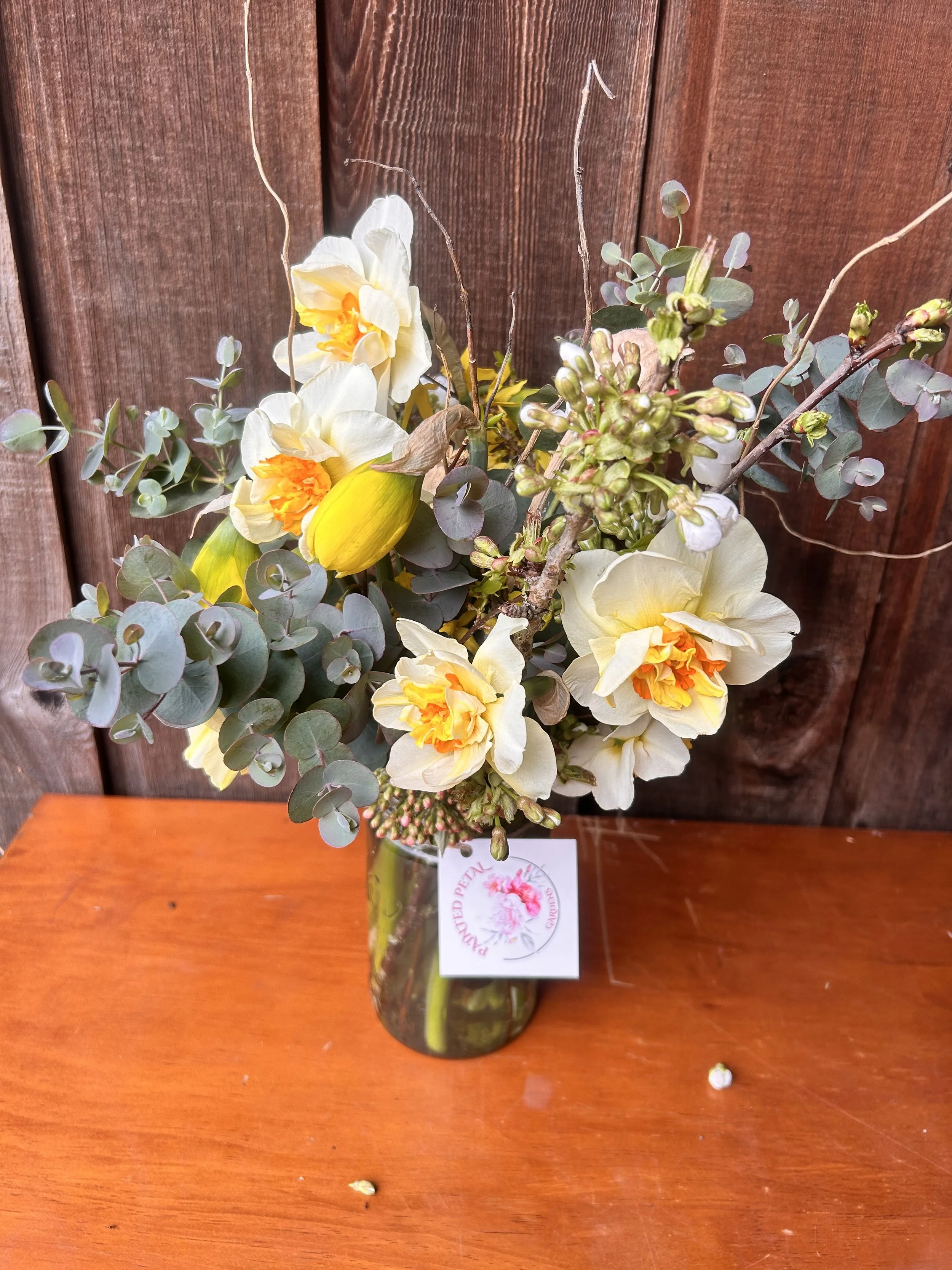 Colorful bouquet of pink, white, and yellow flowers in a white vase on a dark wooden surface against a rustic wooden wall.
