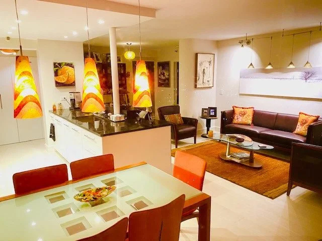 Open-concept living and dining room with a glass-top dining table, orange chairs, a dark recliner, a sofa with orange pillows, a coffee table, and pendant and wall lighting. Kitchen with pendant lights and artwork on walls.
