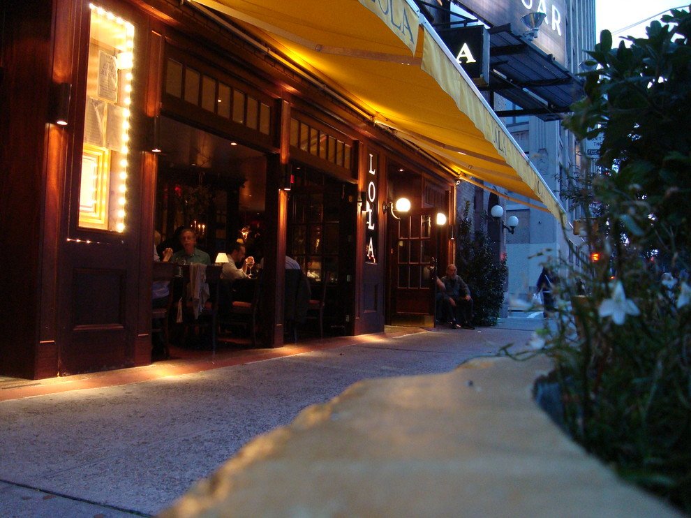 Exterior of a restaurant called 'LOLA' with a yellow awning, illuminated signs, and people dining inside and sitting outside during evening.