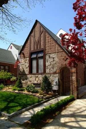 A brick and stone house with a sharply peaked roof, tall windows, and a small front yard with a sidewalk, trees, and bushes.