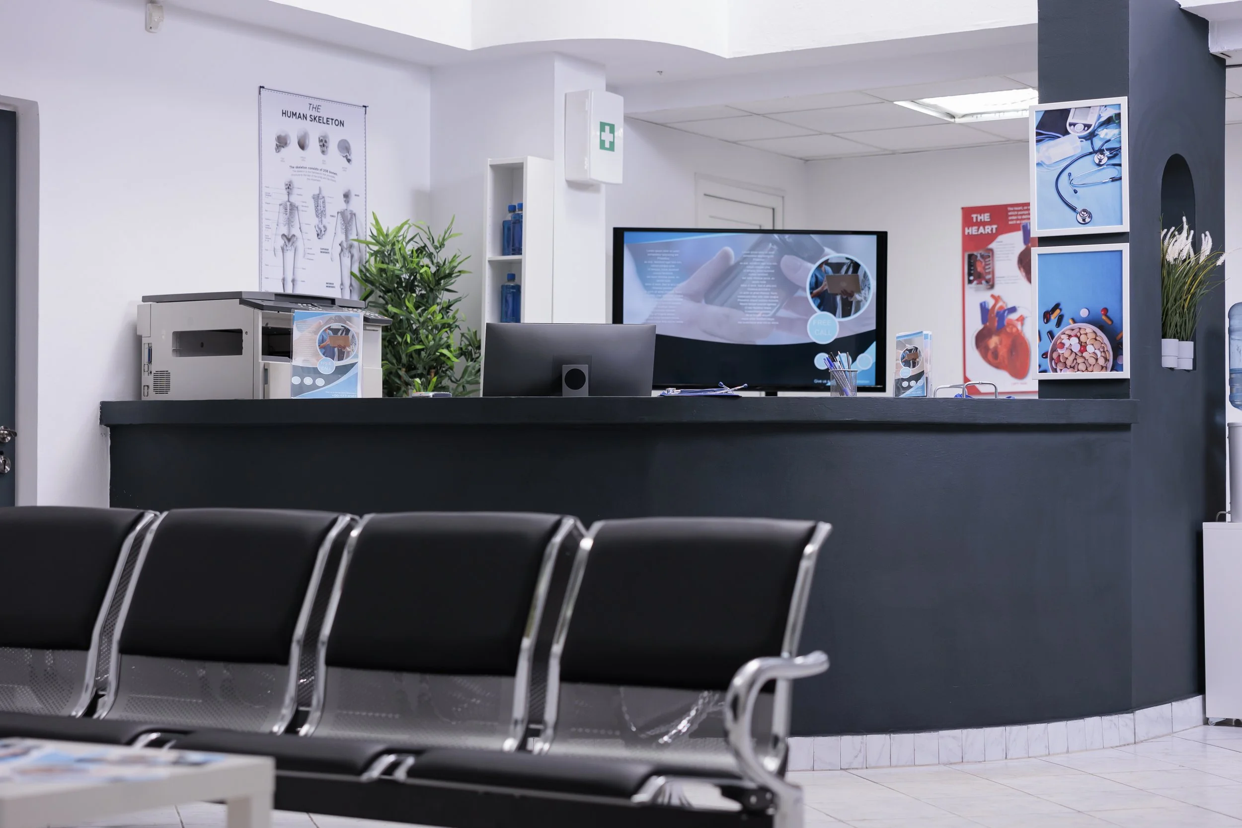 A hospital reception desk with a black countertop, a computer monitor, and a large screen display. Chairs are in the waiting area in front of the desk. Posters about the human skeleton and heart are on the walls, along with a green plant and medical supplies.