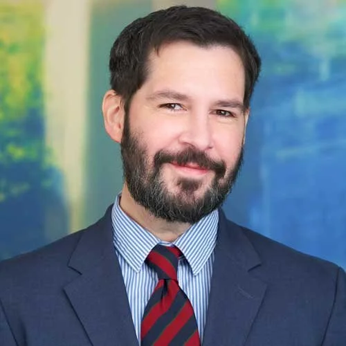 Professional man with dark hair and beard, wearing a navy suit and striped tie, smiling against a colorful abstract background.
