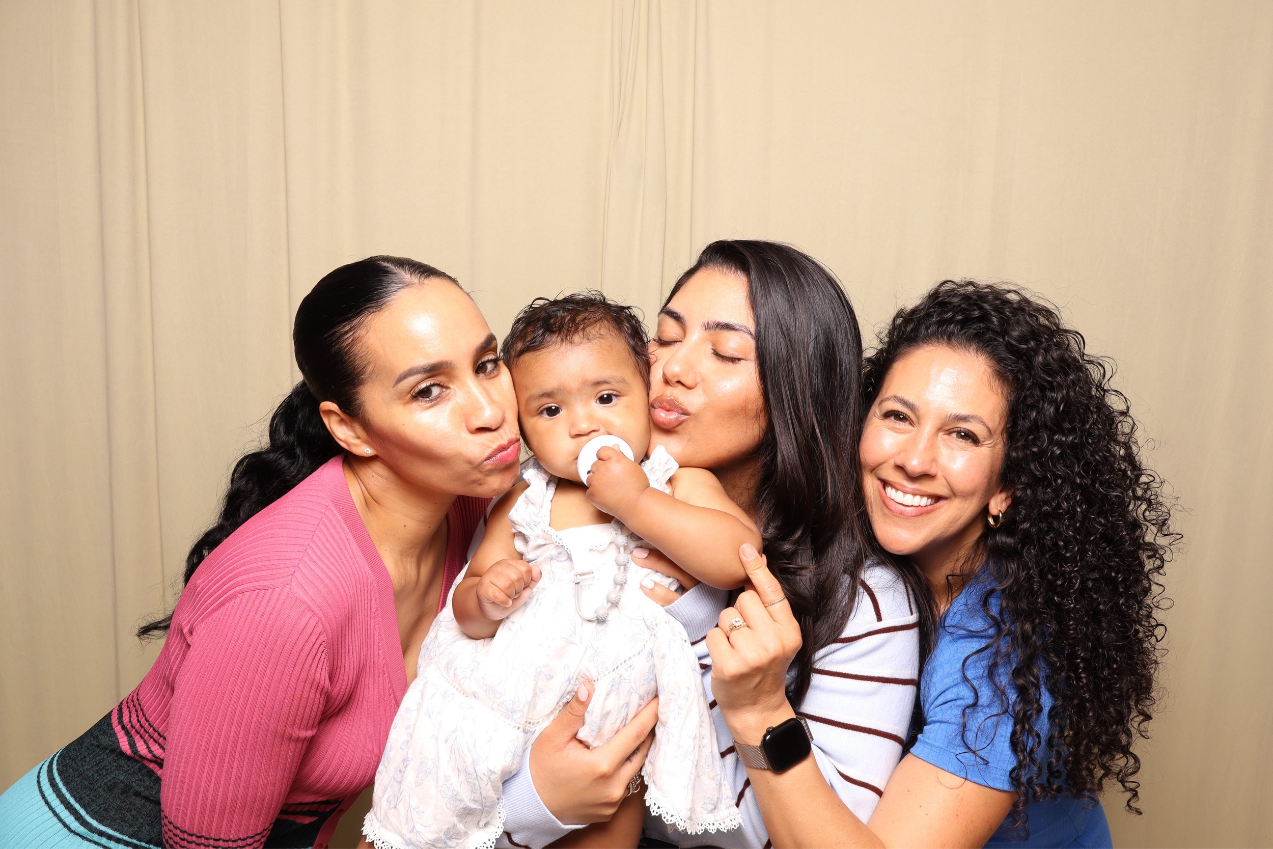 Four women and a young child are gathered closely together, smiling and making kissy faces for the camera against a beige curtain background.