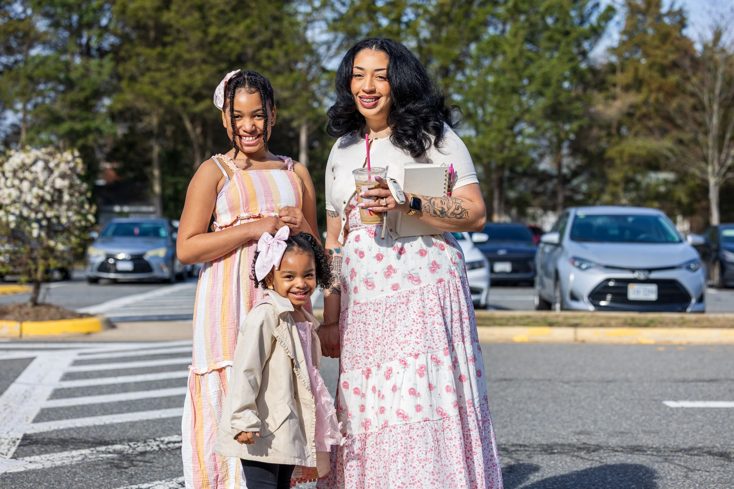A woman and three young girls smiling and standing together in a parking lot with trees and cars in the background. The woman is holding a drink and has tattoos on her arm. The girls are wearing casual, colorful clothing.