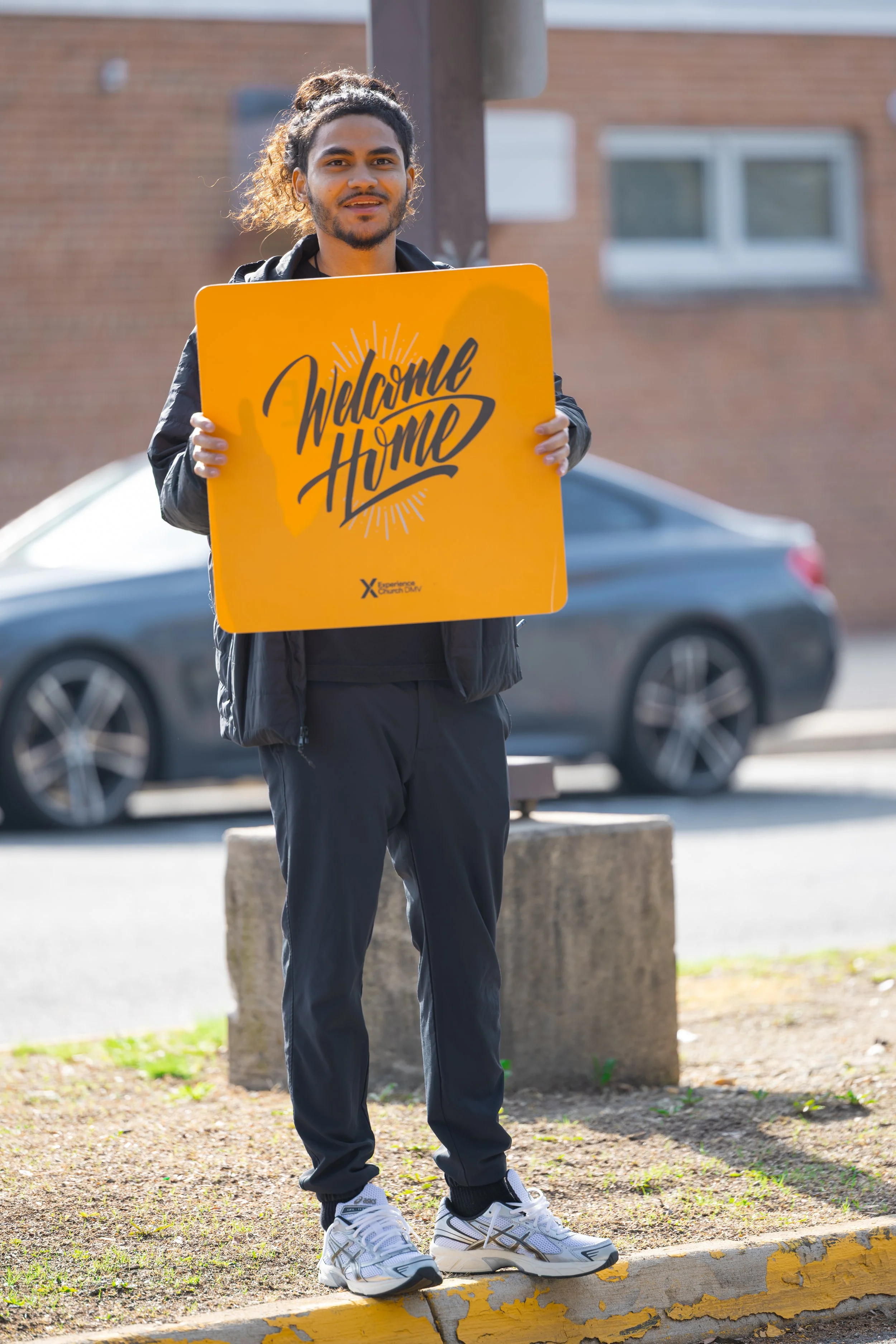 A young man with curly hair holding a yellow 'Welcome Home' sign stands outdoors near a curb and a parking lot, with a brick building and a gray car in the background.