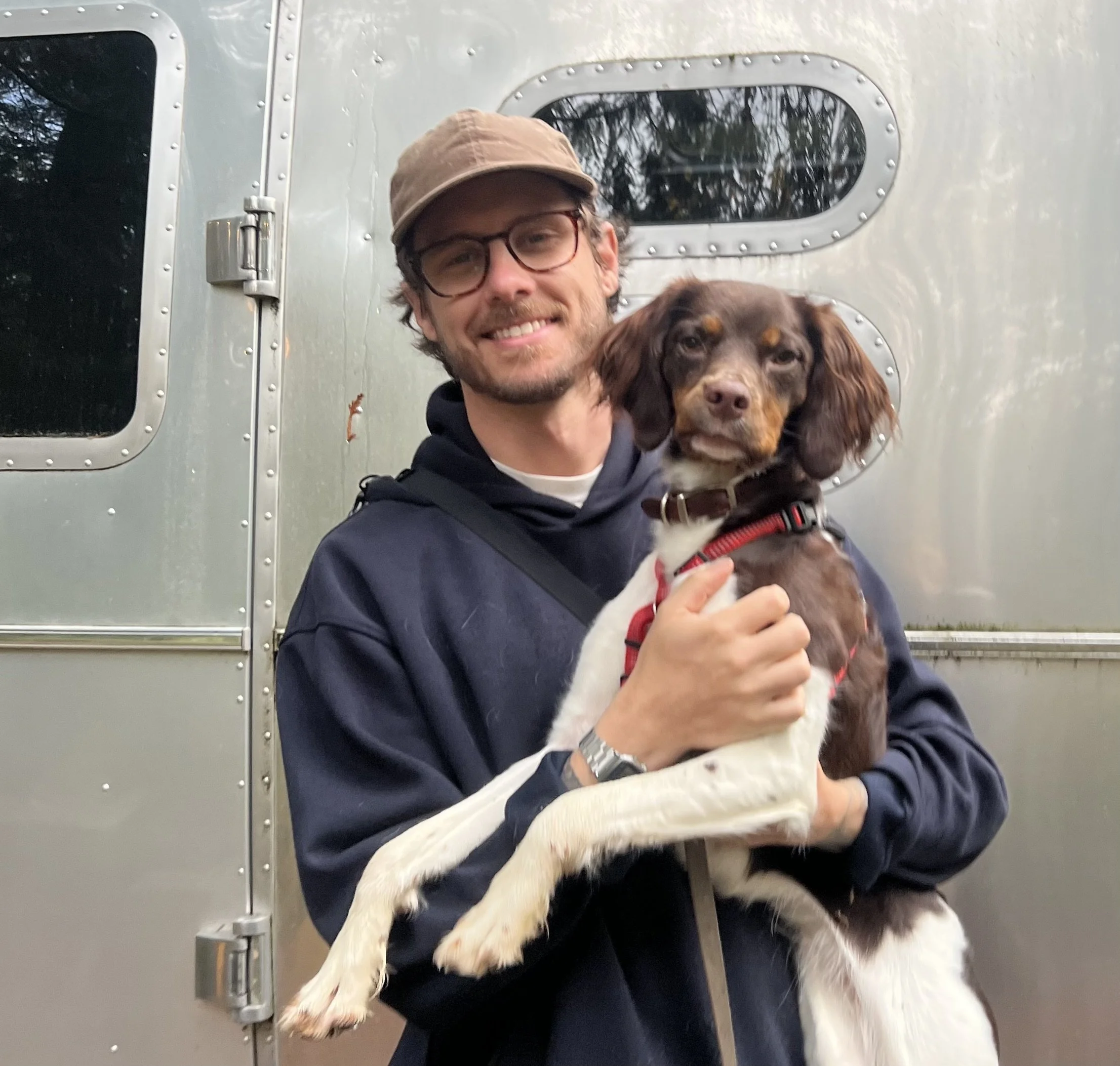 A man with glasses and a brown cap holds a brown and white dog in front of a silver trailer.