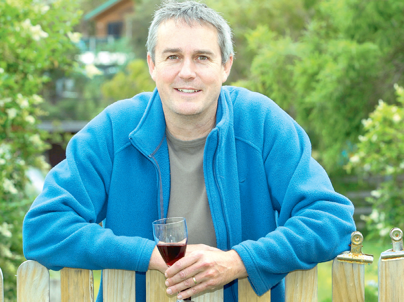 A middle-aged man with short gray hair and blue eyes, smiling, sits outdoors holding a glass of red wine with both hands, wearing a blue fleece jacket and a gray t-shirt, with a blurred green garden and trees in the background.
