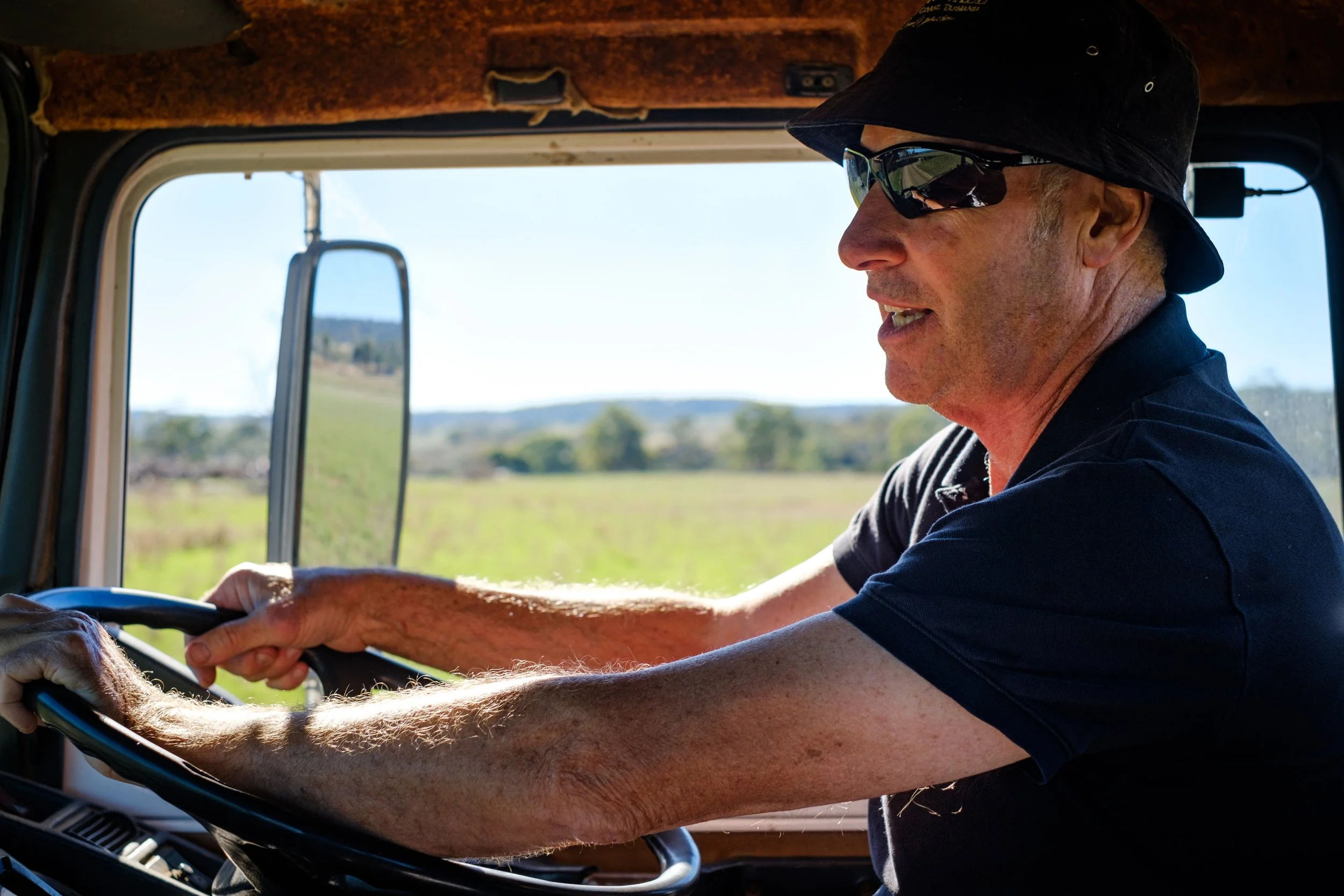 Rodney Lyne driving a large truck in a rural area, wearing sunglasses and a black hat, with a scenic landscape visible through the window.