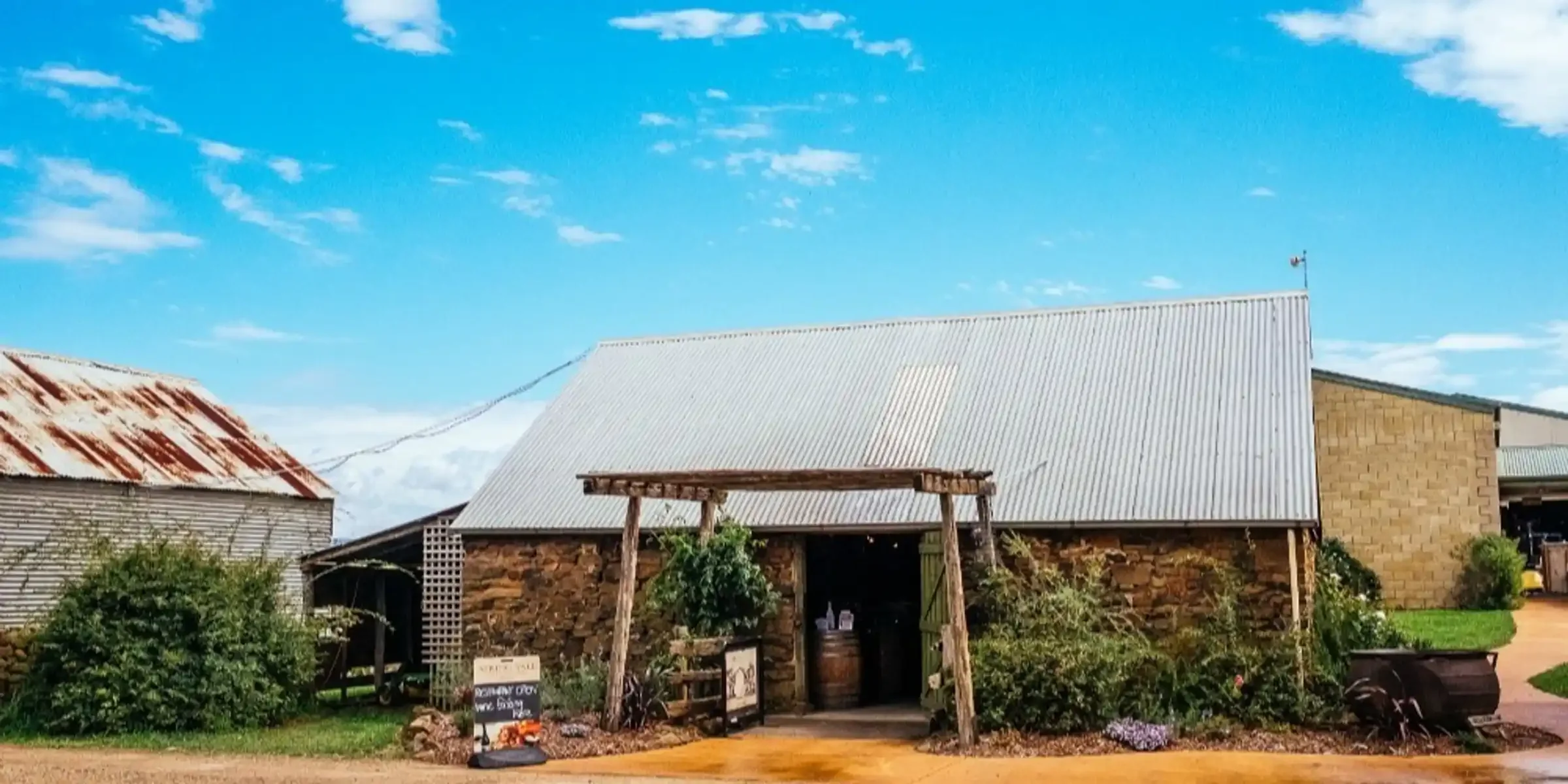 A rustic farm building with a corrugated metal roof, a stone and brick facade, surrounded by greenery and a dirt pathway, under a bright blue sky with scattered clouds.
