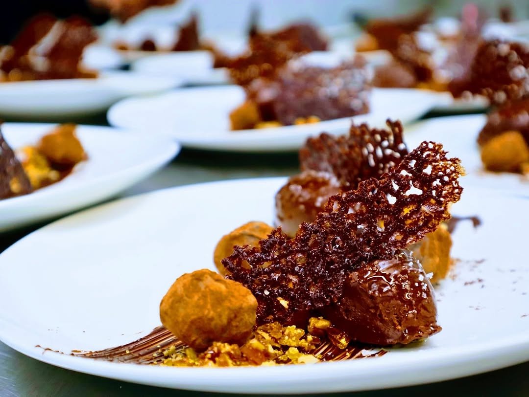 Multiple white plates with gourmet desserts, including chocolate, caramel, and crunchy toppings, arranged on a table.