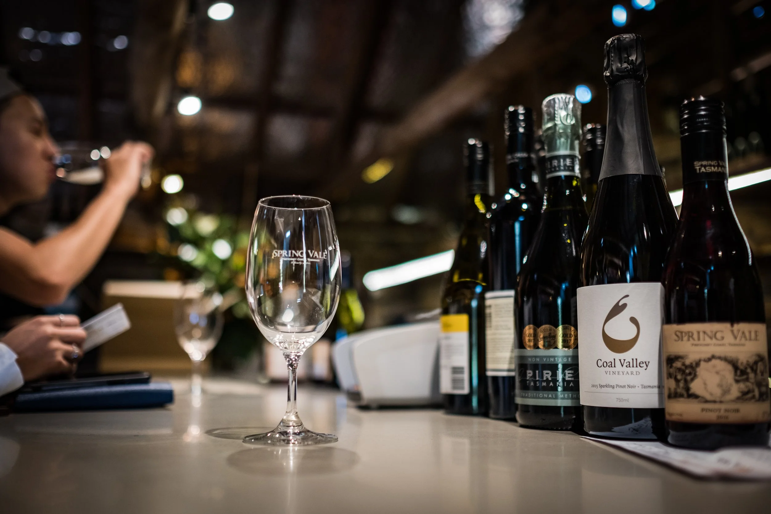 Empty wine glass on the Spring Vale bar counter with several wine bottles in the background and a person drinking from a glass in the blurred background.