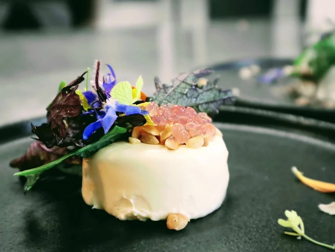 A plated gourmet dish with a creamy white base topped with pinkish-orange caviar and garnished with edible flowers, herbs, and leaves.