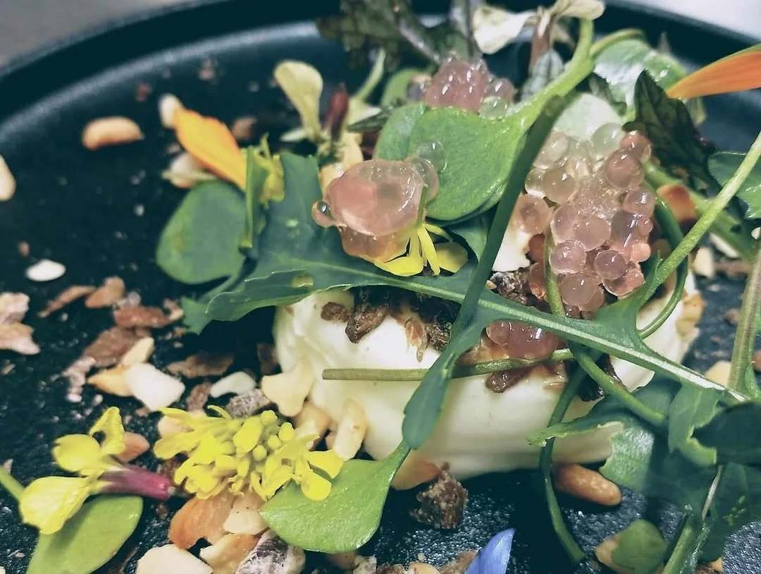Close-up of a dome-shaped appetizer topped with green leaves, pinkish translucent tapioca pearls, and garnished with edible flowers and nuts on a black plate.