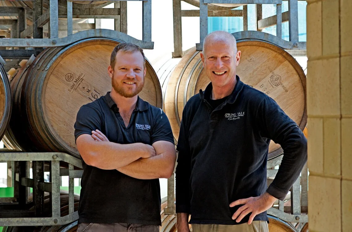 Rodney & Tim Lyne standing in front of stacked wine barrels at a winery, smiling and wearing black polo shirts with the Spring Vale WInes logo.