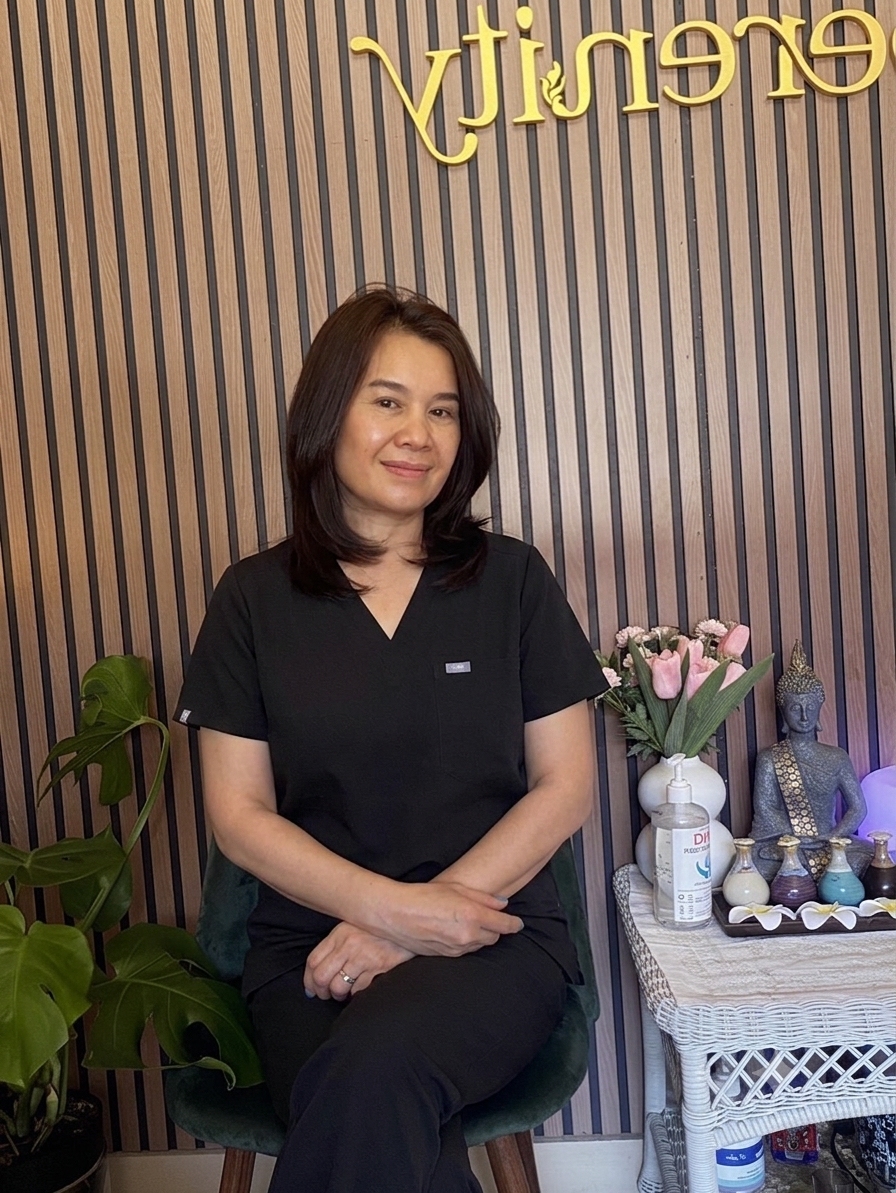 Professional female massage therapist in grey uniform sitting in a warm Thai spa room with candles and plants, representing Serenity Thai Wellness & Massage in Los Angeles.