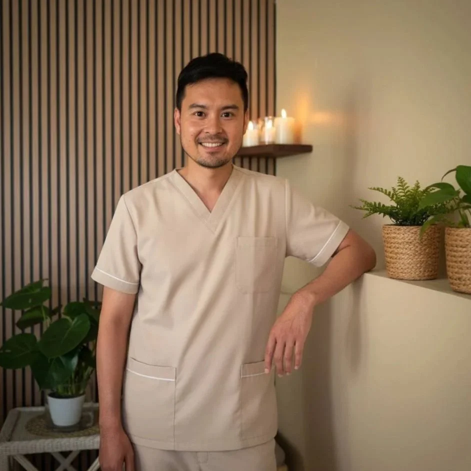 Professional male massage therapist in grey uniform standing in a warm Thai spa room with candles and wooden wall, representing Serenity Thai Wellness & Massage in Los Angeles.