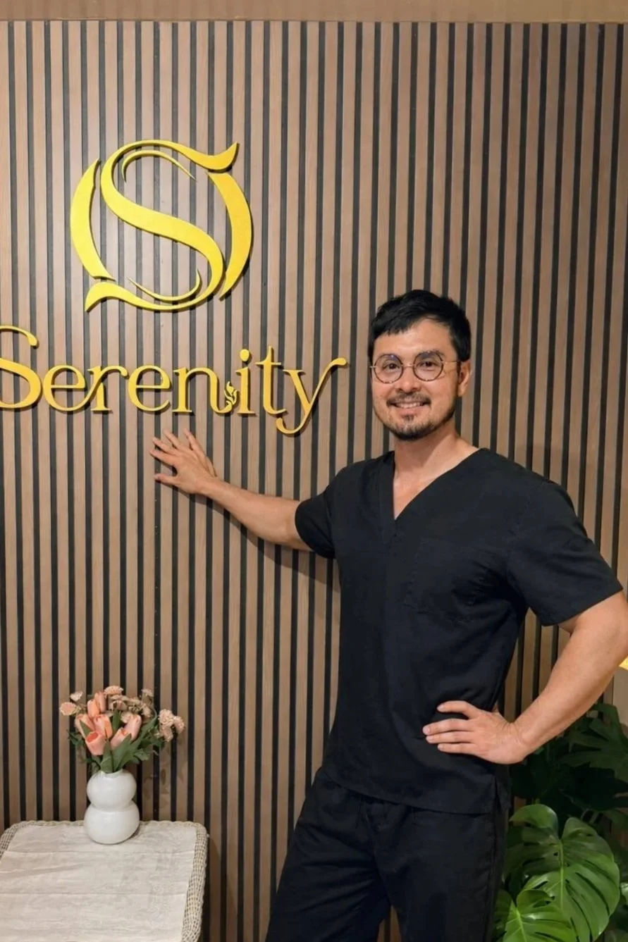 Professional male massage therapist in grey uniform standing in a warm Thai spa room with candles and wooden wall, representing Serenity Thai Wellness & Massage in Los Angeles.