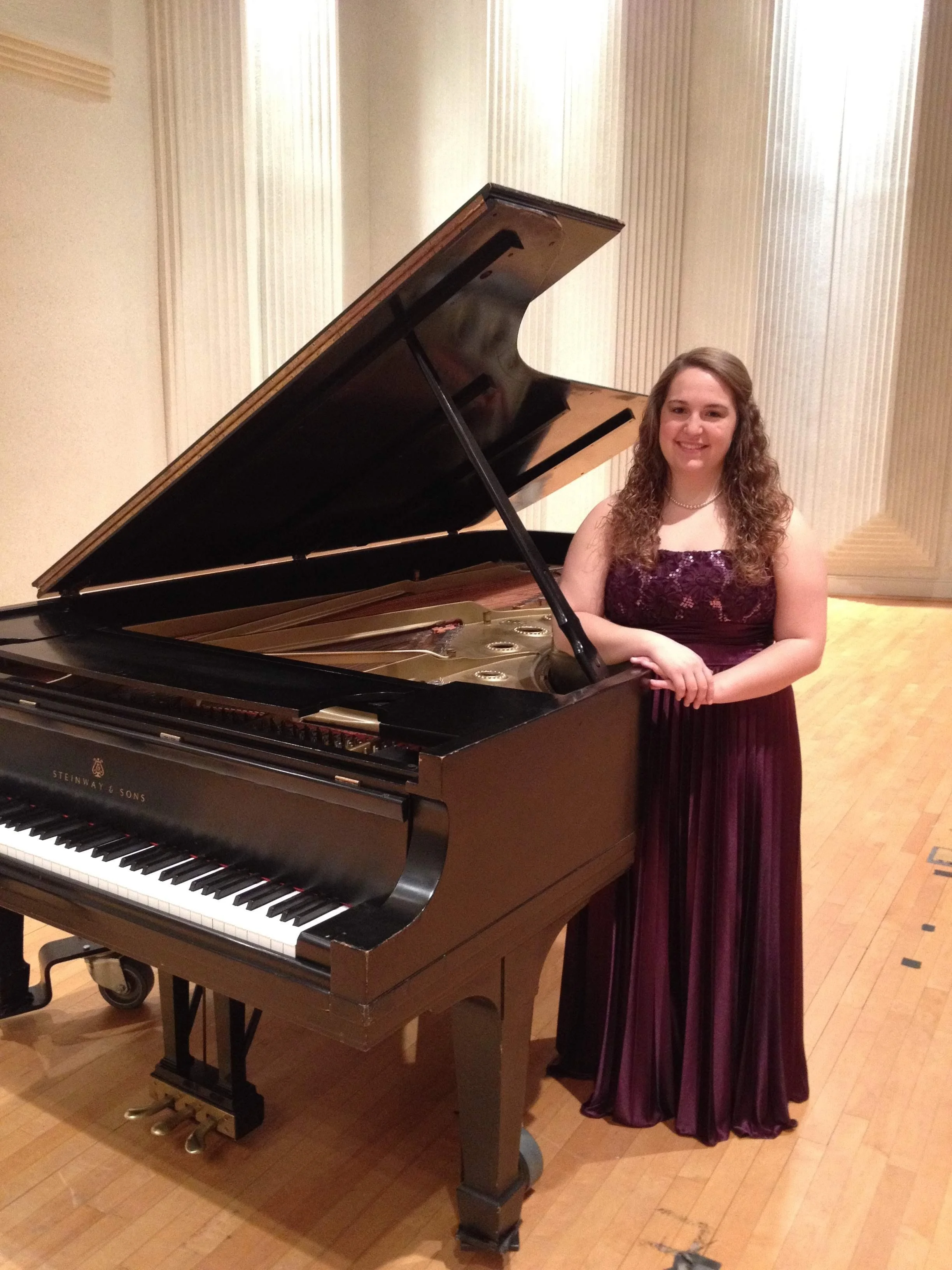 A young woman in a burgundy evening gown standing next to a grand piano in a music hall.