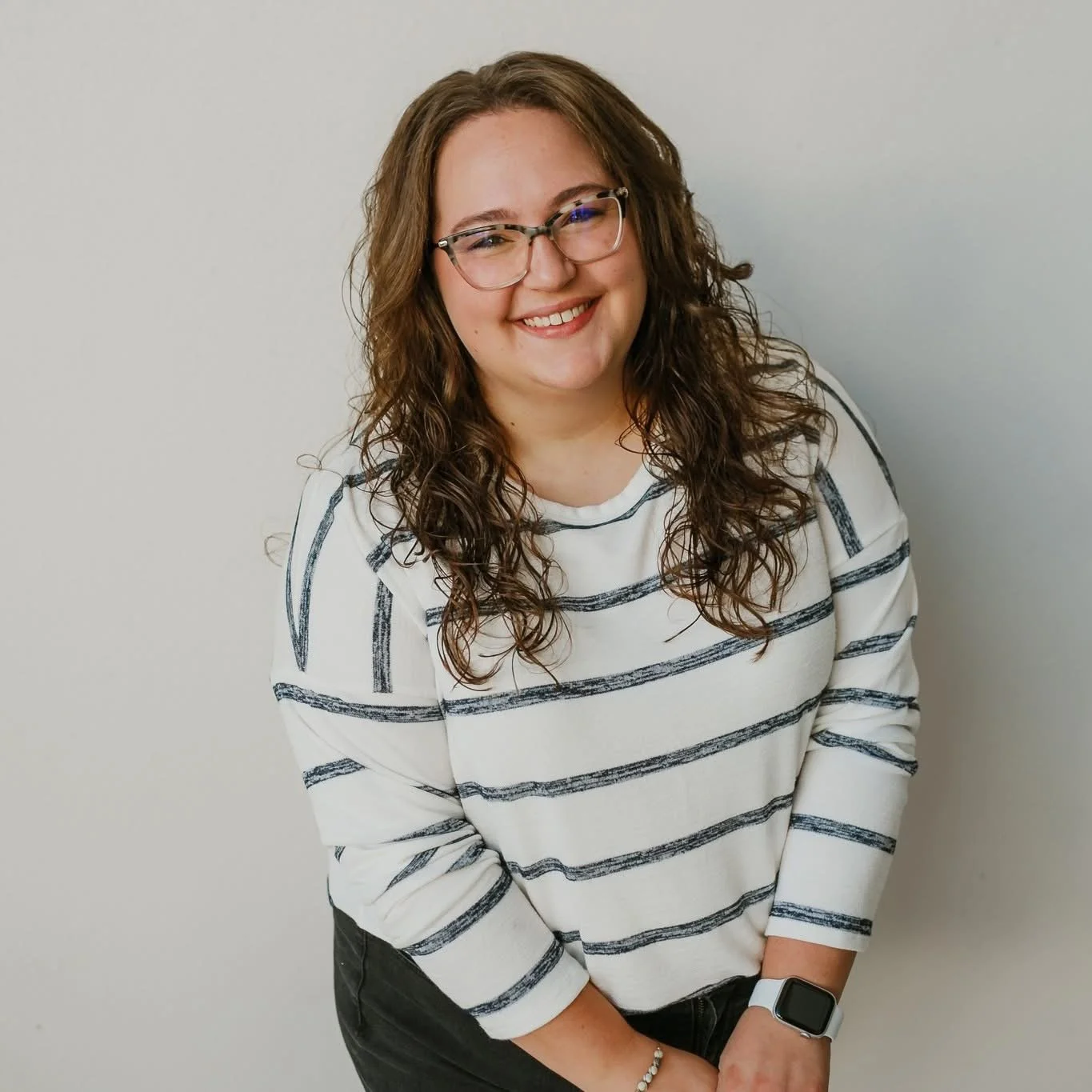A woman with curly brown hair, glasses, and a watch, smiling and standing against a plain light-colored wall, wearing a striped long-sleeve shirt.