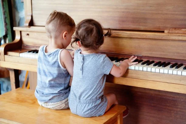 Two young children sitting on a wooden bench, playing a piano together in a cozy indoor setting.