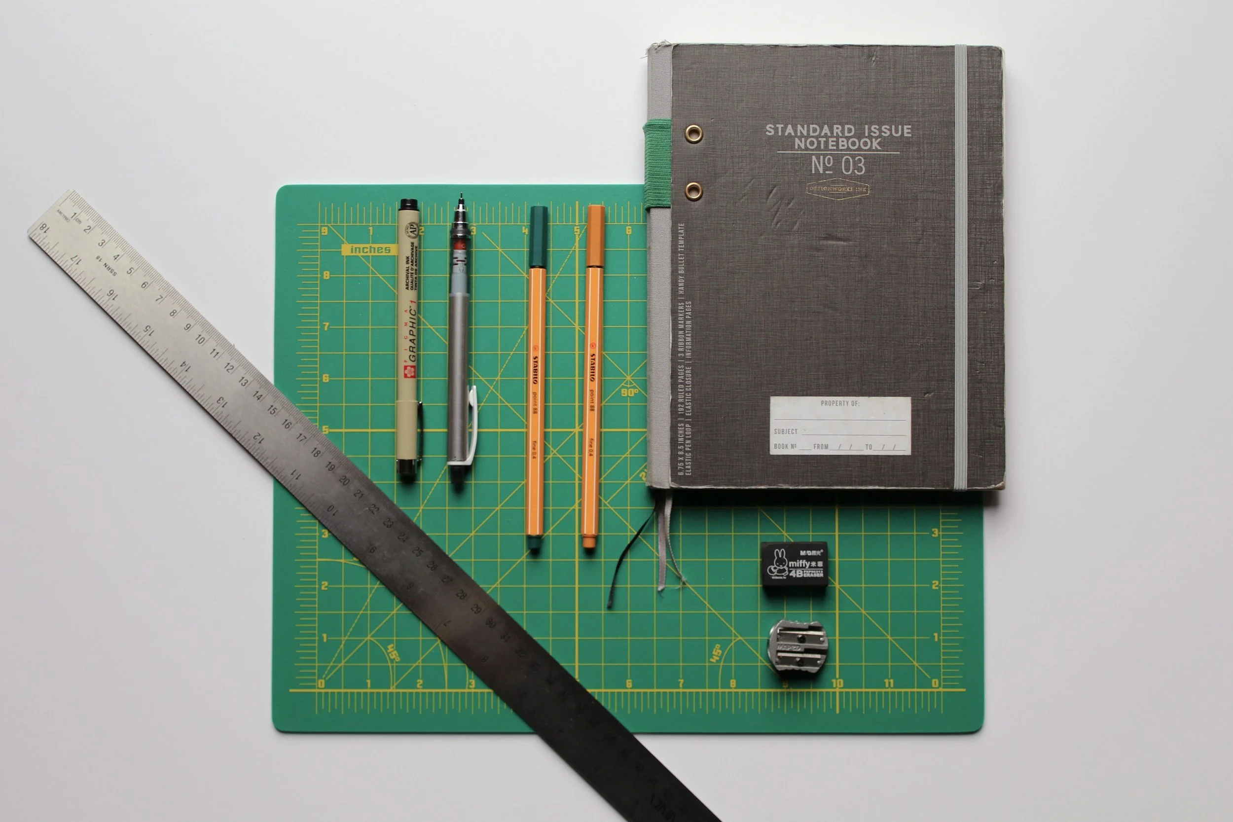 A green cutting mat with rulers, behind which are several pens, a notebook, a small eraser, and a pencil sharpener.