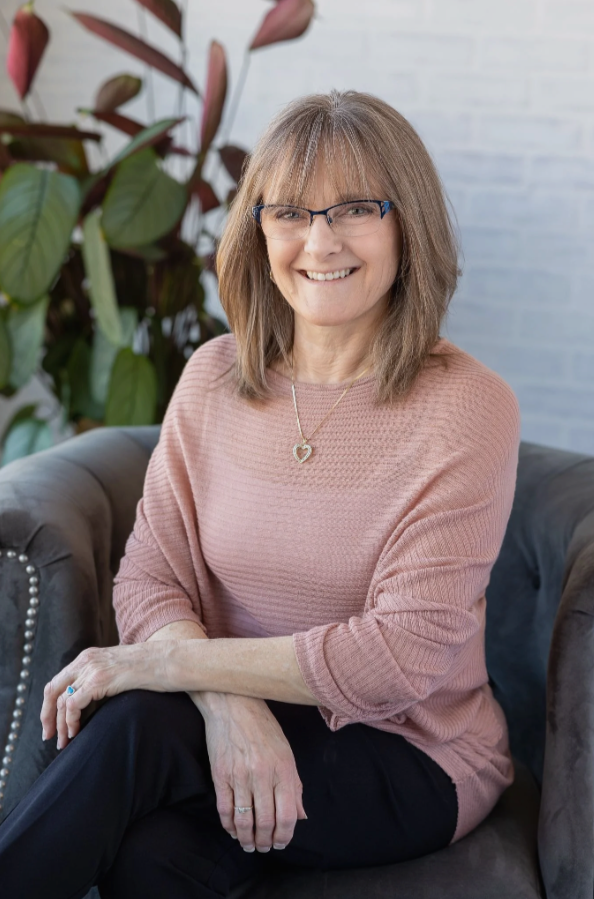 A smiling middle-aged woman with shoulder-length light brown hair, wearing glasses, a pink sweater, and a heart-shaped necklace, sitting on a dark gray armchair indoors. There are green plants and a white brick wall in the background.