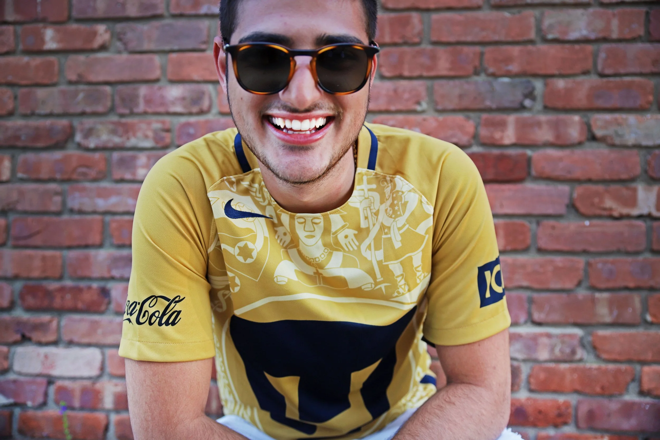 A young man wearing sunglasses and a yellow sports jersey with black and white designs, smiling in front of a brick wall.