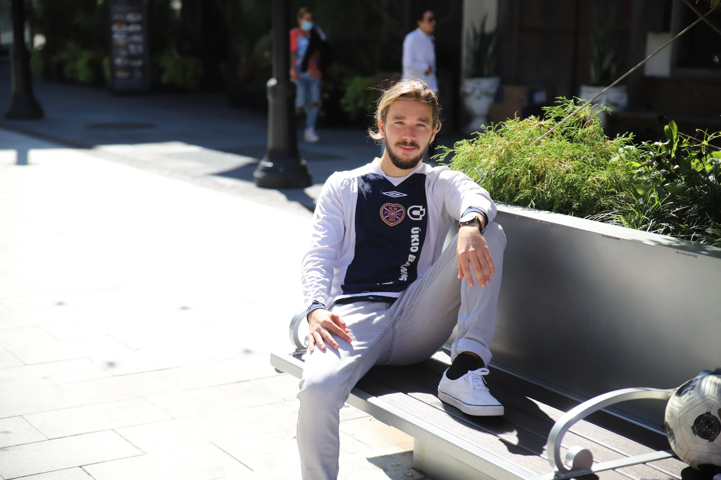 Young man with light brown hair, beard, wearing a white and navy blue soccer jersey with a crest, sitting casually on a park bench in bright sunlight, with a soccer ball on the ground beside him.