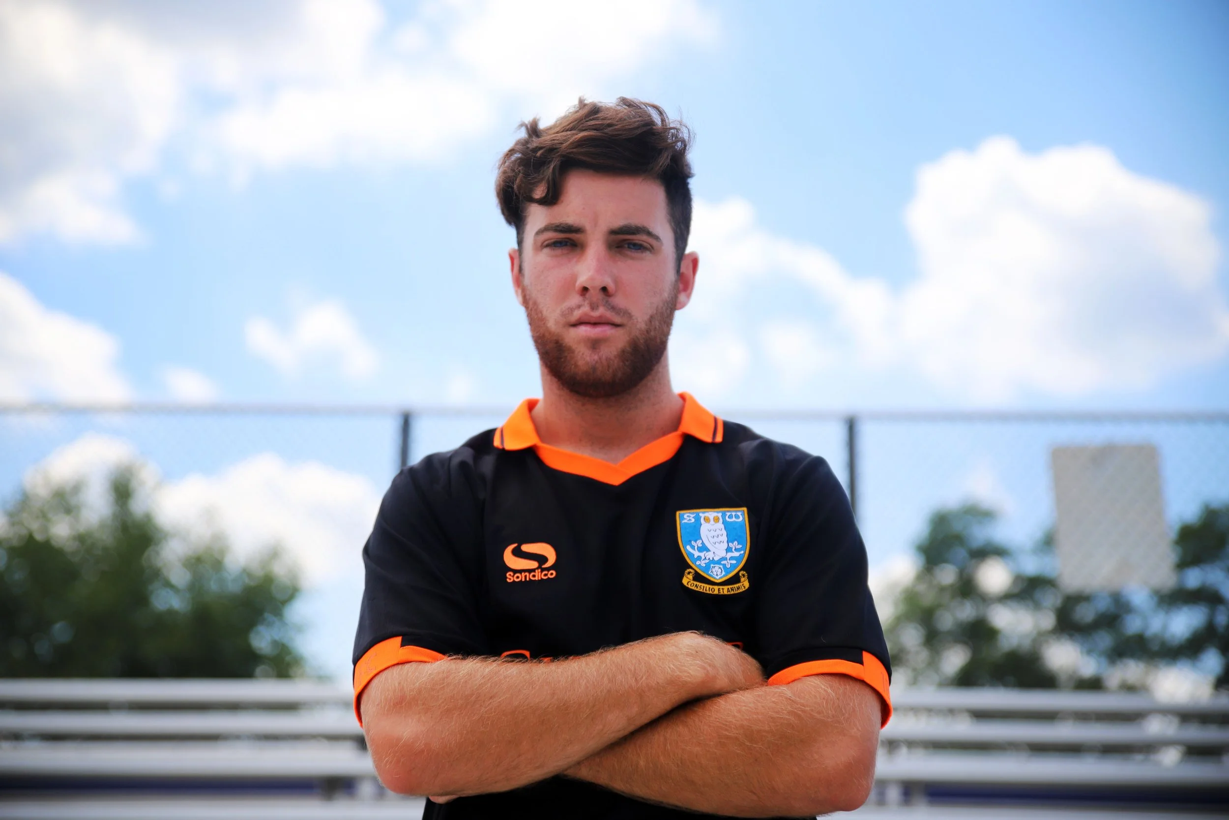 A young man stands outdoors with arms crossed, wearing a black sports jersey with orange accents and a crest, against a blue sky with white clouds and stadium bleachers in the background.