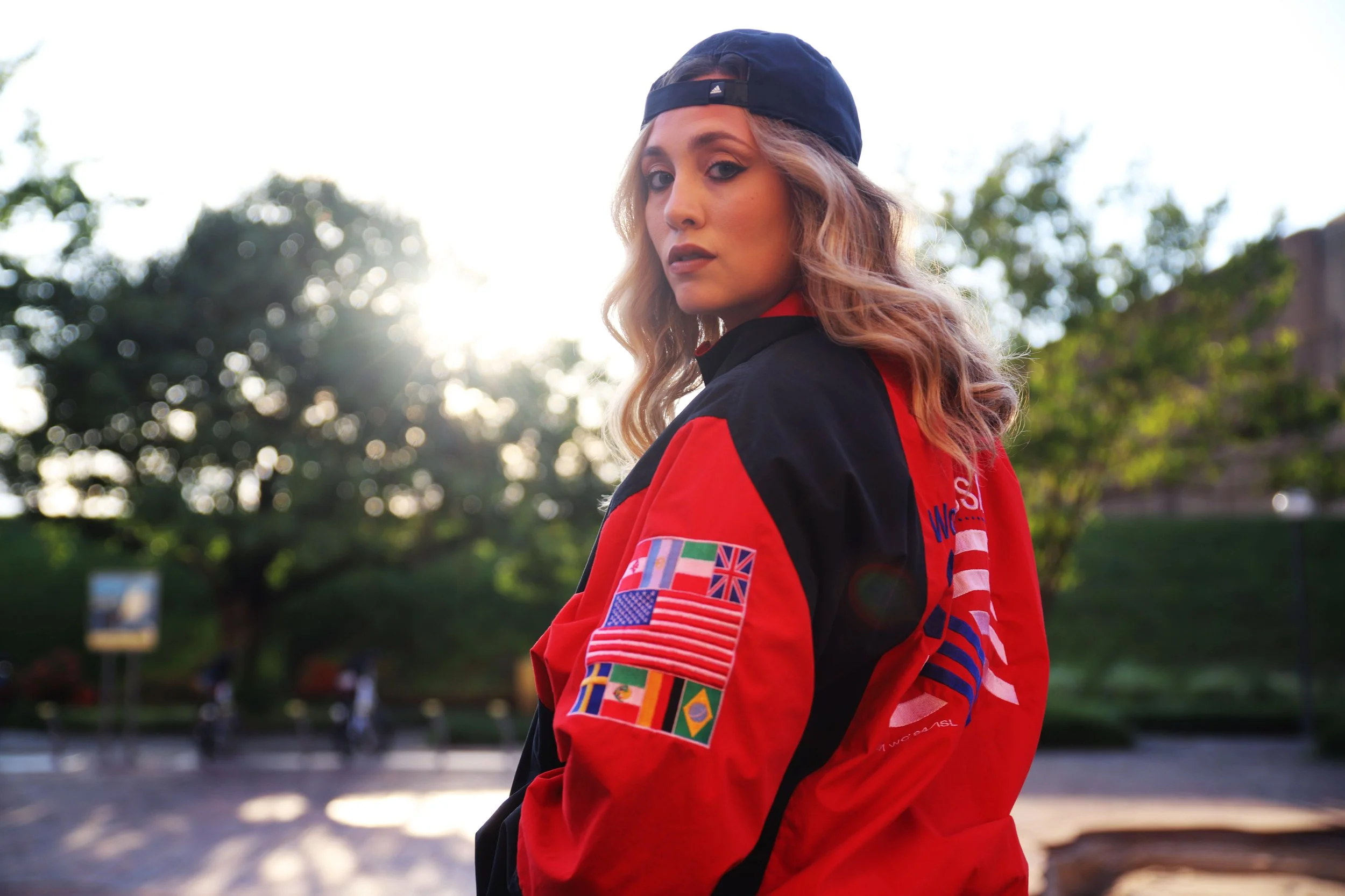 Young woman with wavy blond hair wearing a black and red jacket with patches, including flags of various countries, and a black baseball cap turned backward, standing outdoors with trees and sunlight in the background.