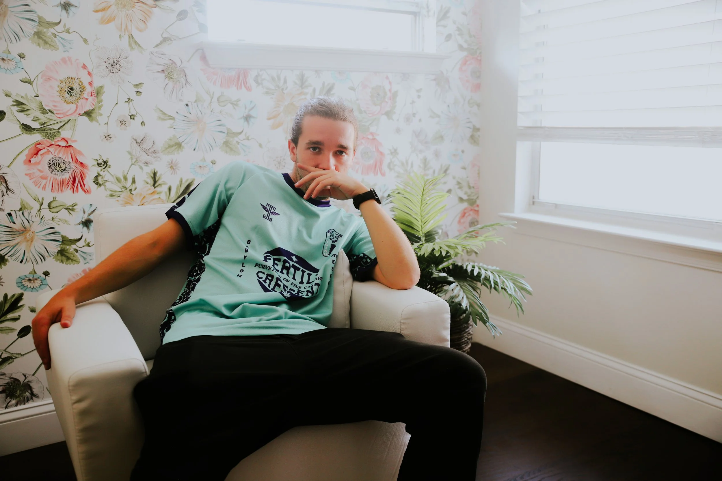 A young man with long hair tied back, wearing a mint green sports jersey and black pants, sitting on a white armchair in a bright room with floral wallpaper and blinds on the window. He is resting his left arm on the armrest and covering part of his 