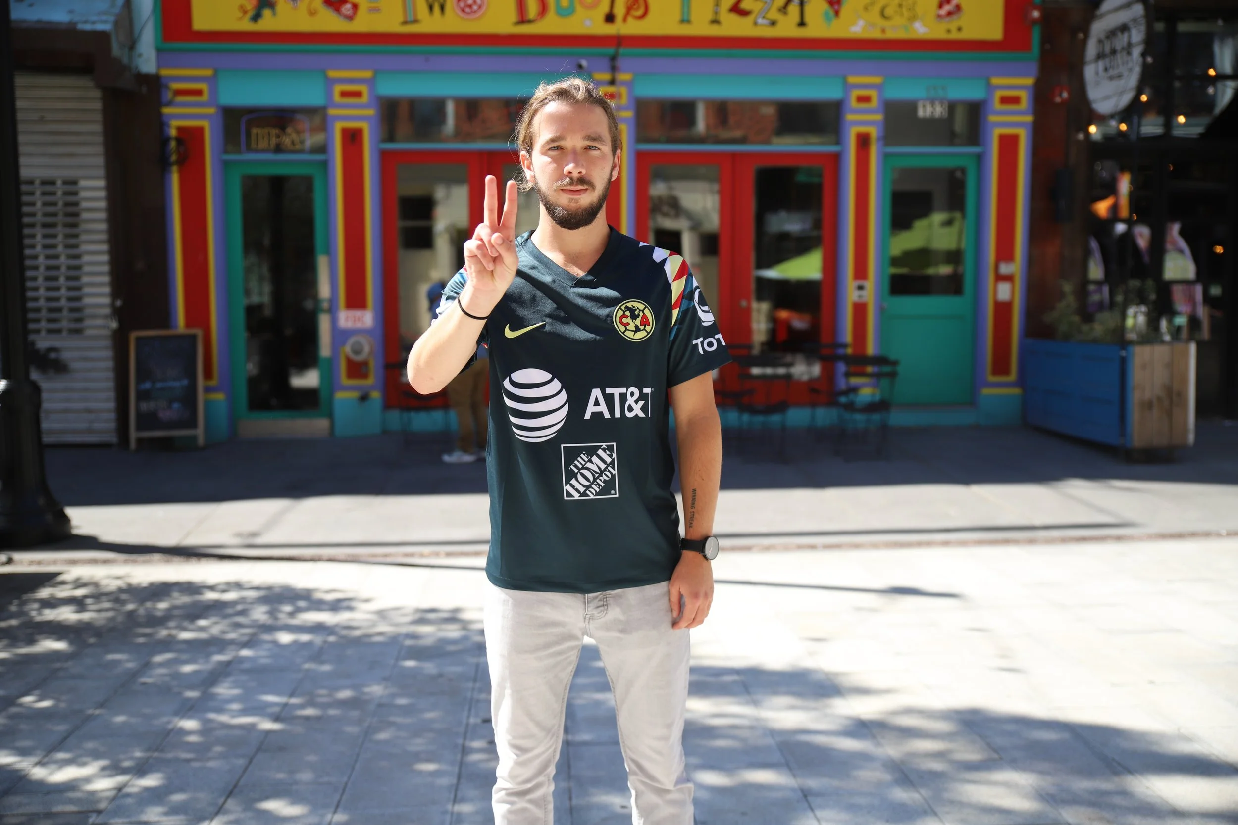 A young man with a beard and long hair tied back, wearing a black soccer jersey with sponsor logos, making a peace sign with his right hand, standing outside in front of a colorful restaurant or store with a bright yellow, blue, and red painted facad