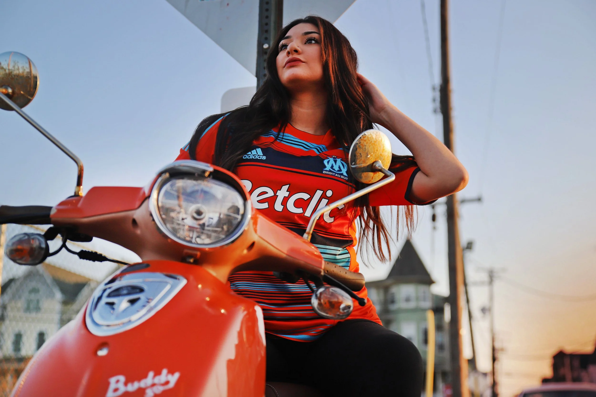 A woman with long dark hair sitting on an orange scooter, wearing a red and blue soccer jersey, with a sunset sky and telephone poles in the background.