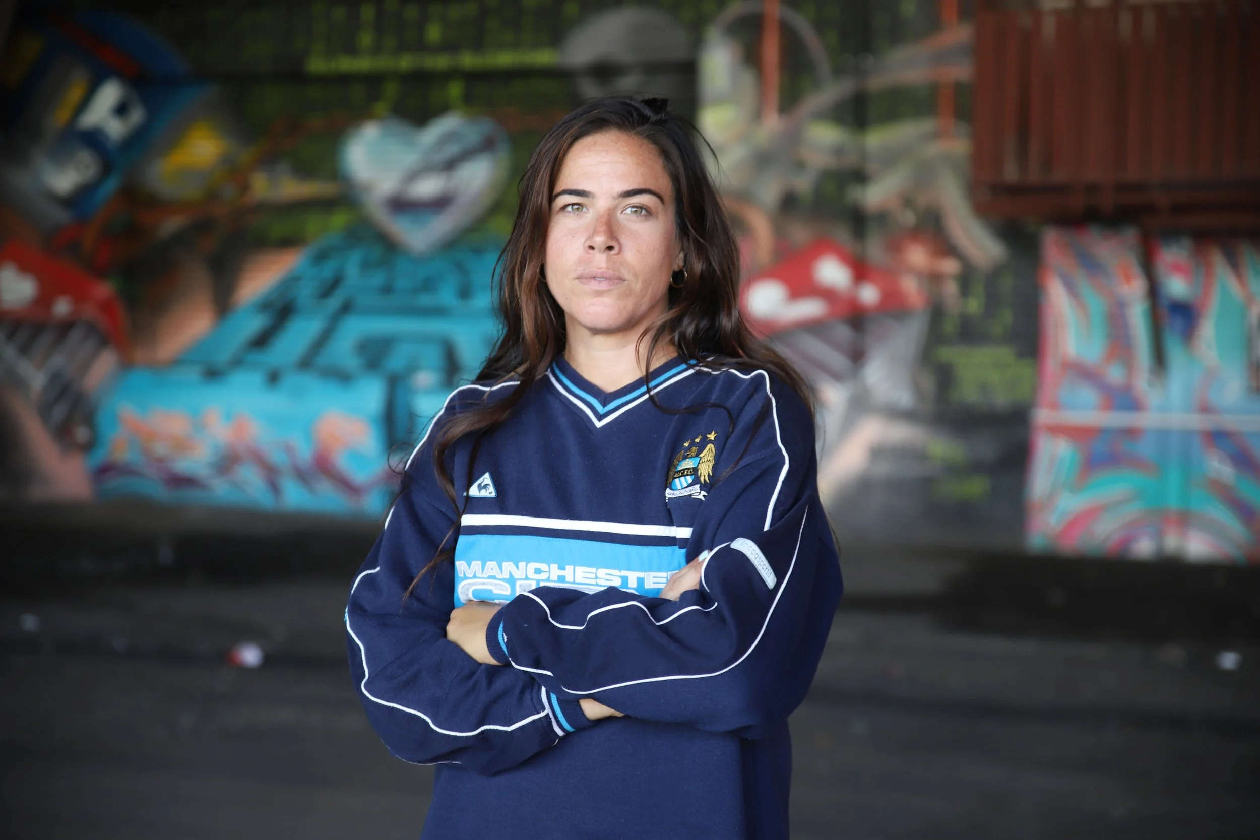 A woman standing with arms crossed wearing a Manchester City football jersey in front of a graffitied wall.