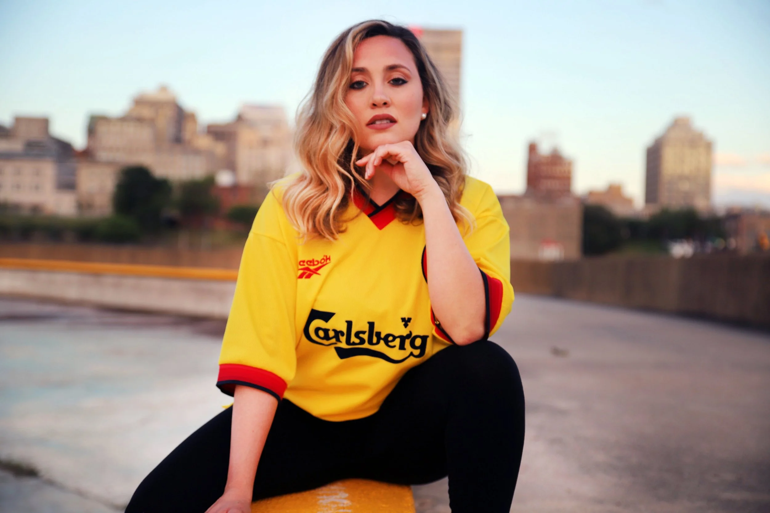 Woman with wavy blonde hair wearing a yellow sports jersey with Carlsberg logo, sitting on concrete surface with cityscape in background during sunset.