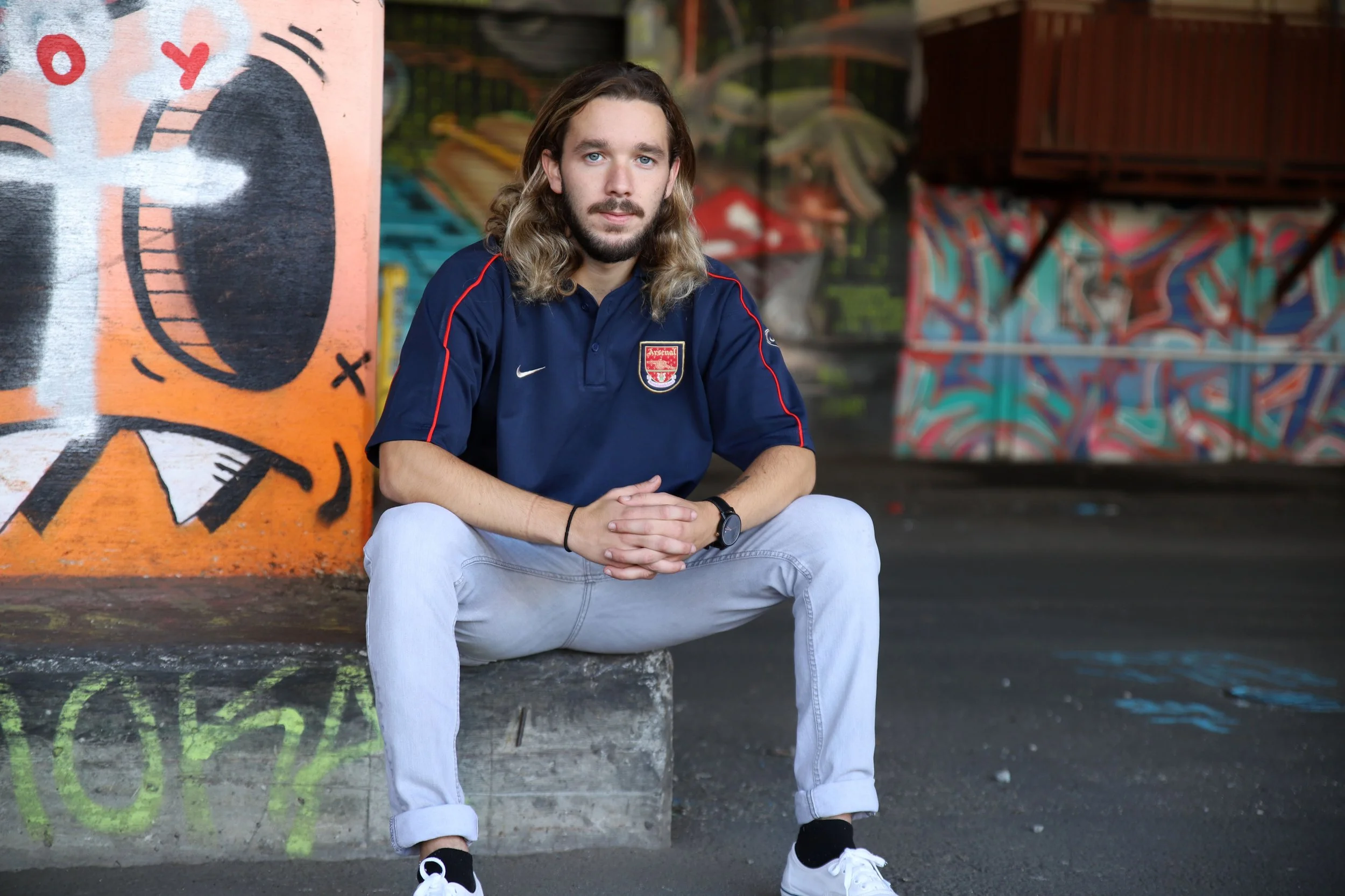 Young man with long hair and beard sitting on a concrete block in front of colorful street art graffiti, wearing a navy Arsenal football club jersey, light jeans, and white sneakers.