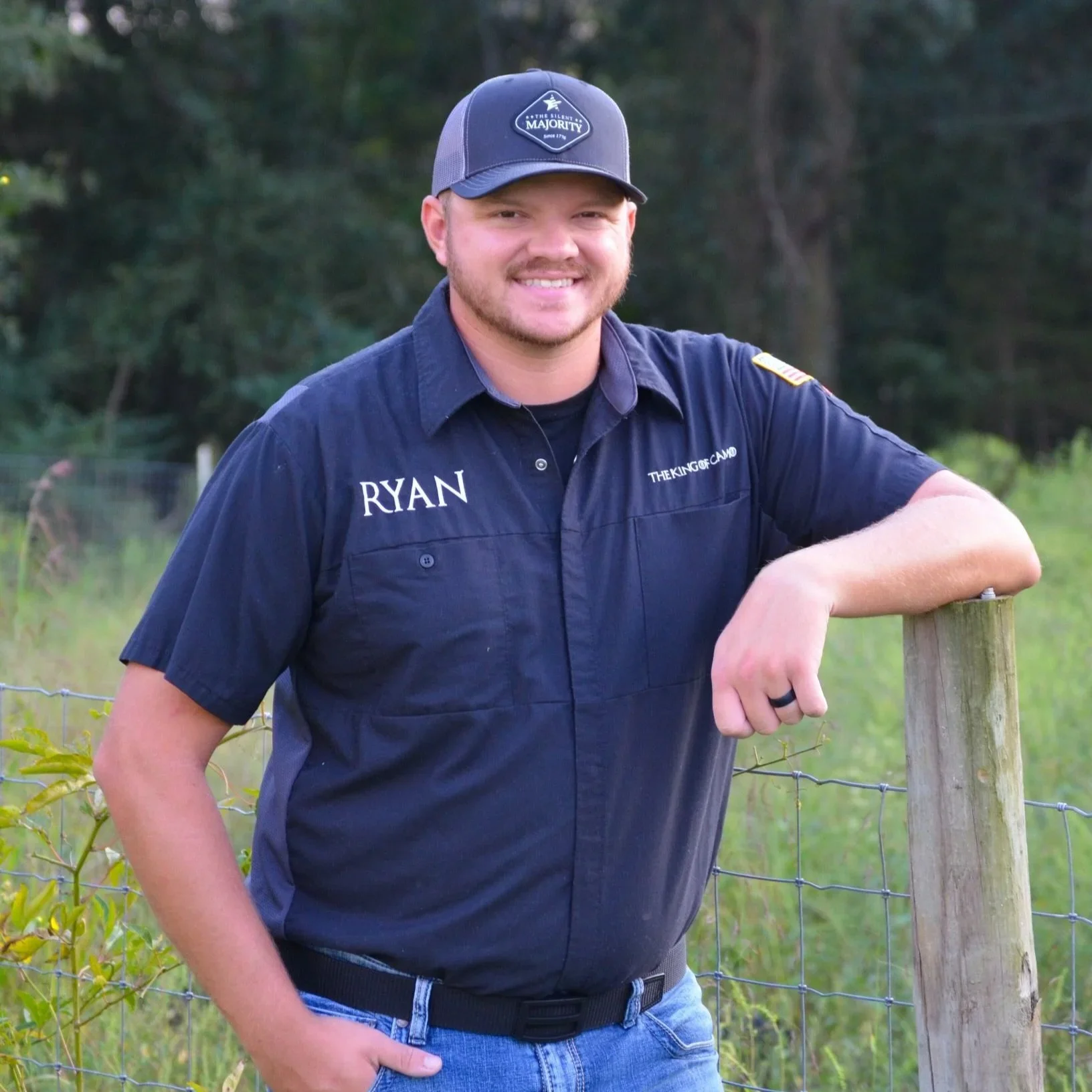 A man wearing a navy blue shirt with 'RYAN' embroidered on it, standing outdoors near a wooden fence, smiling at the camera.