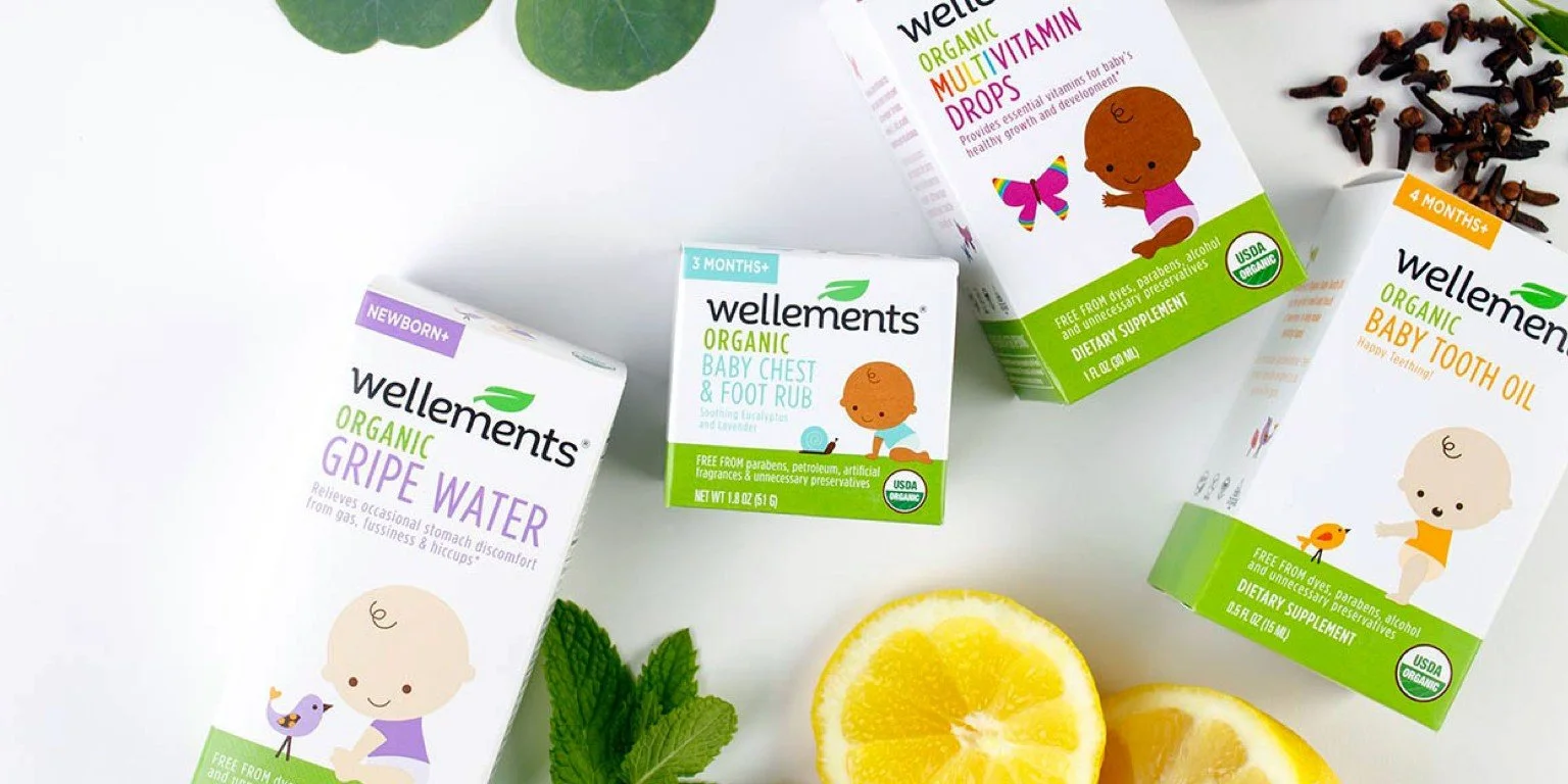 Wellements organic baby care products, including diaper rash cream, baby chest and foot rub, multivitamin drops, and baby tooth oil, with lemon slices, mint leaves, and a few cloves around them.