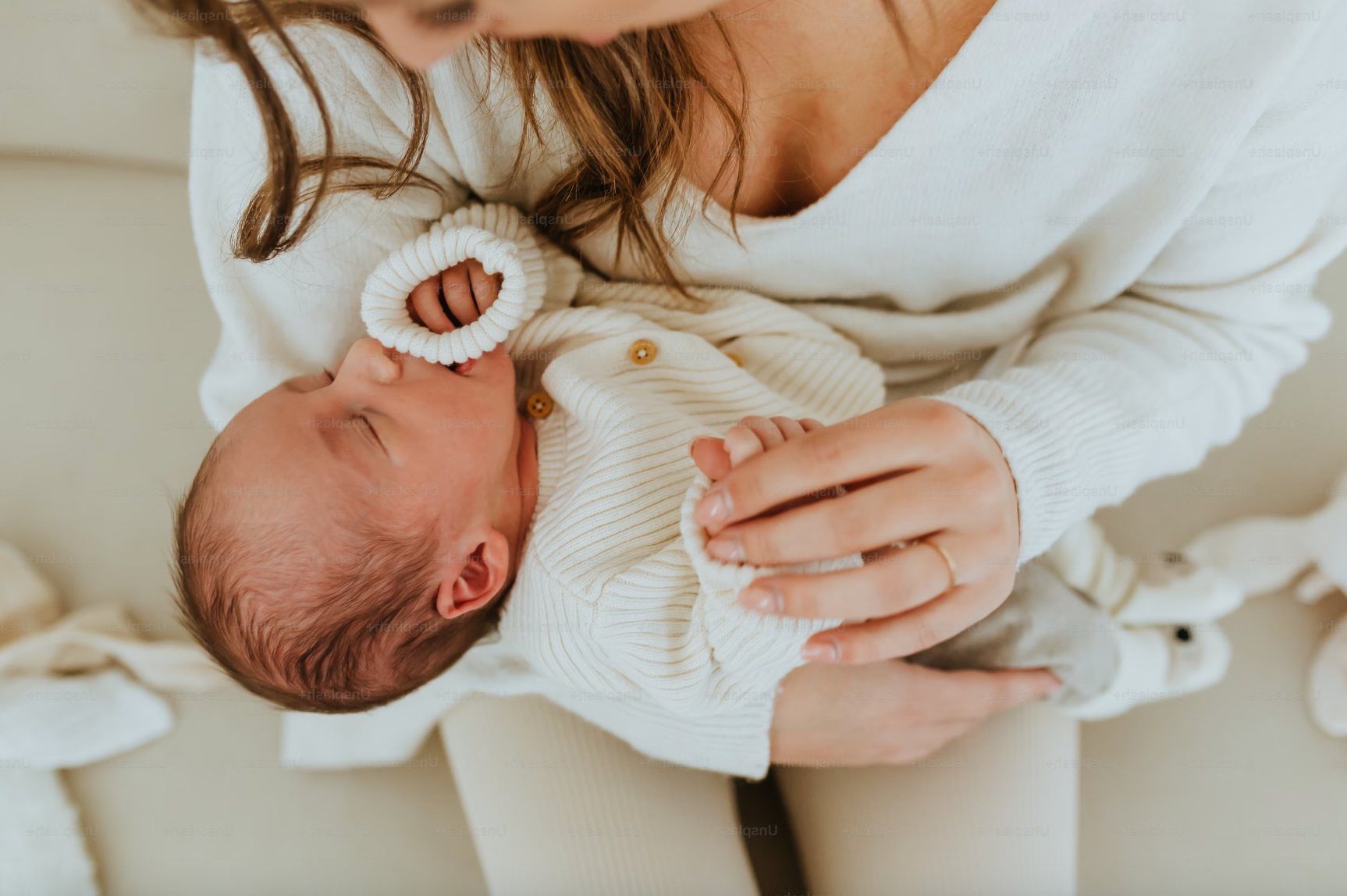 A woman holding a newborn baby, both dressed in cream-colored knit clothing. The woman is gently cradling the baby, who is sleeping peacefully.