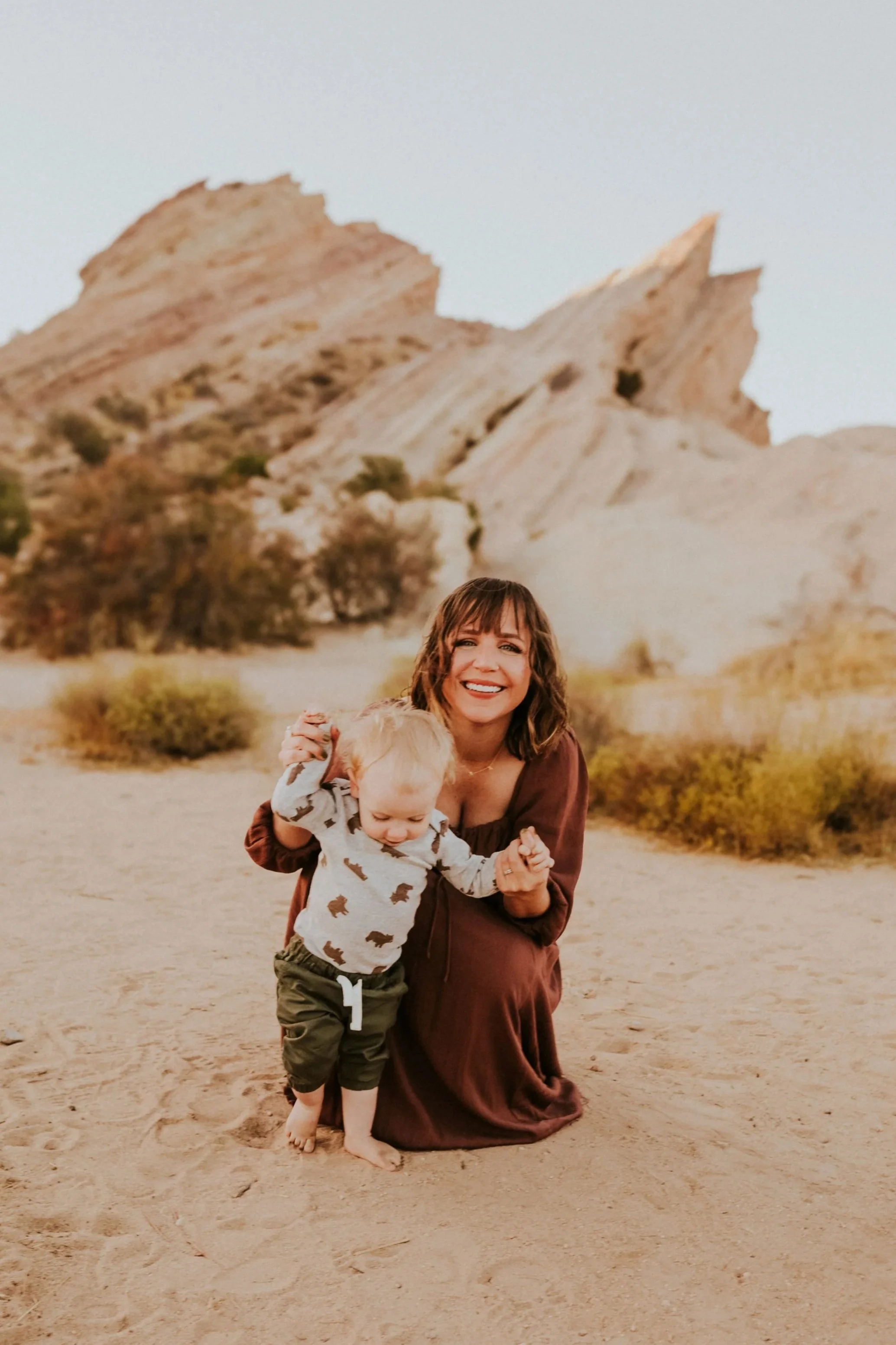 A woman and a young child smiling and playing on a sandy terrain with desert-like rock formations in the background.